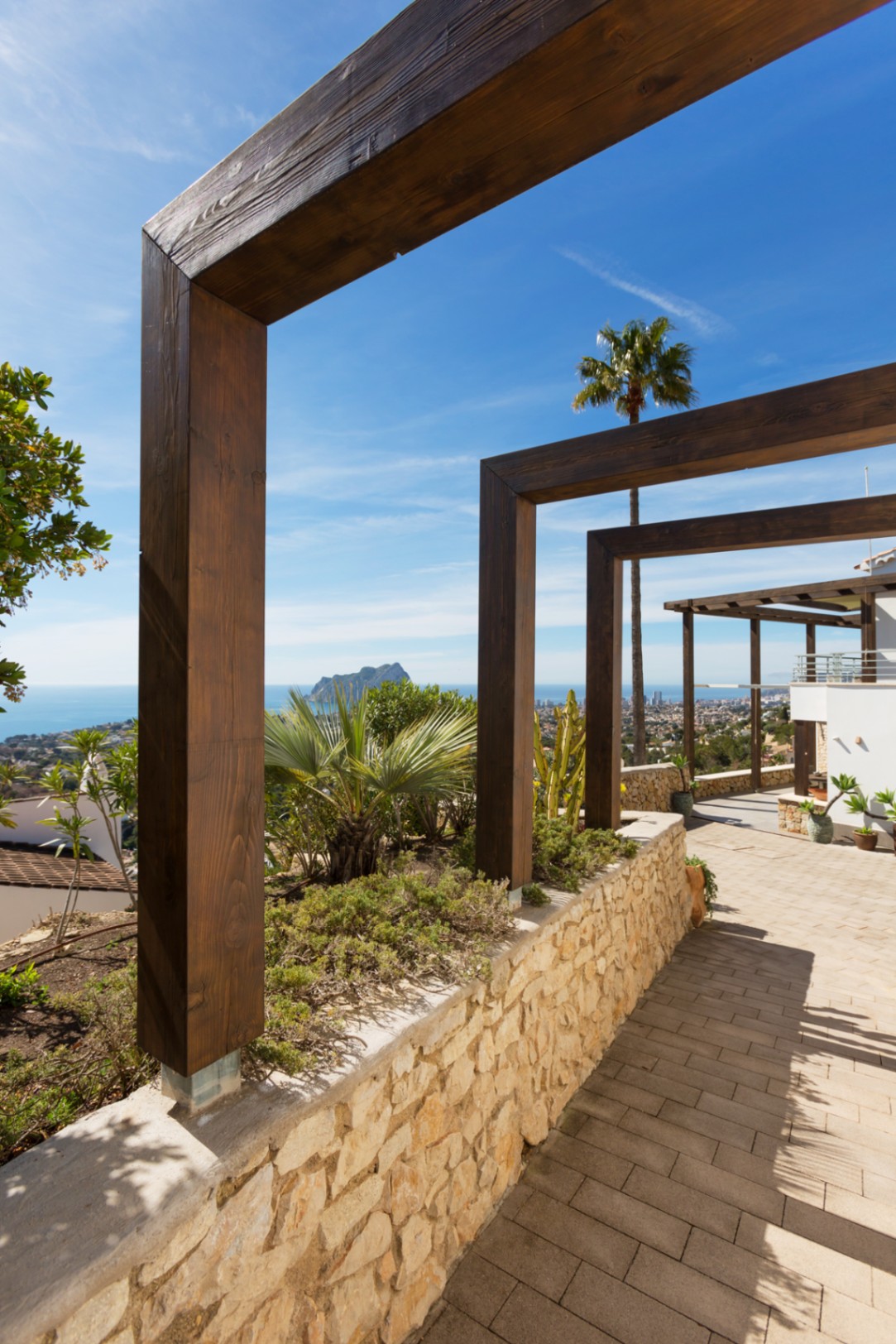 Außenweg mit dunklen Holzpergolen, Natursteinmauer und Panoramablick auf das Mittelmeer und den Peñón de Ifach in der Ferne.