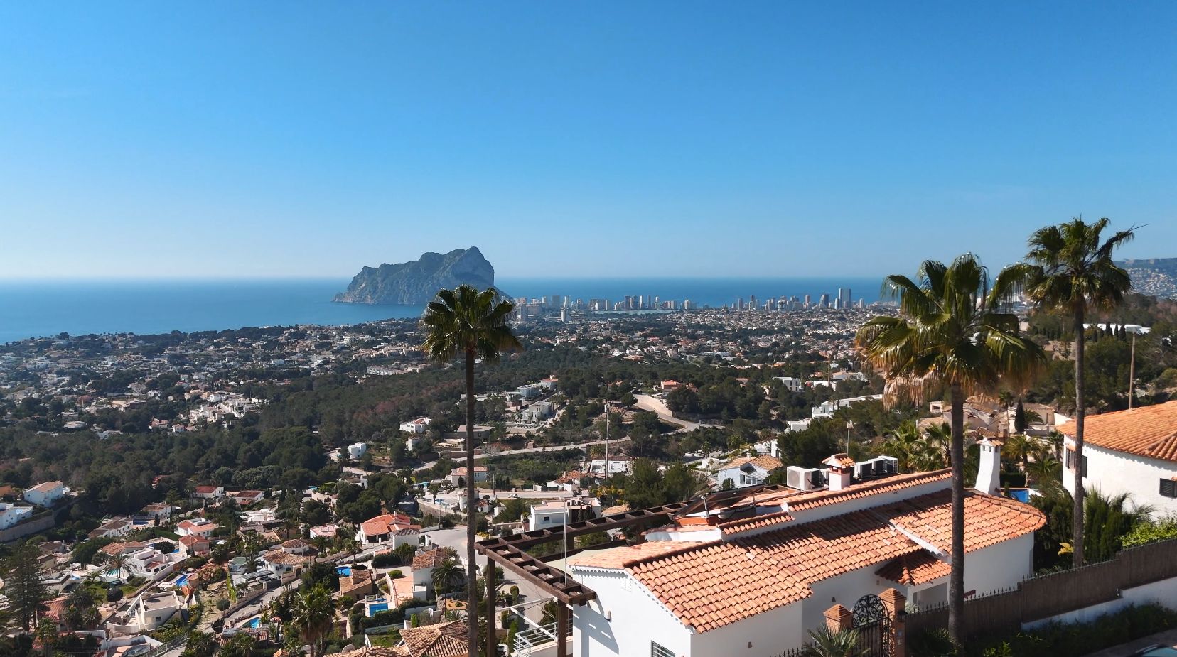 Erhöhter Panoramablick auf den Peñón de Ifach und die Skyline von Calpe, mit mediterranen Villen, Terrakottadächern und Palmen im Vordergrund.