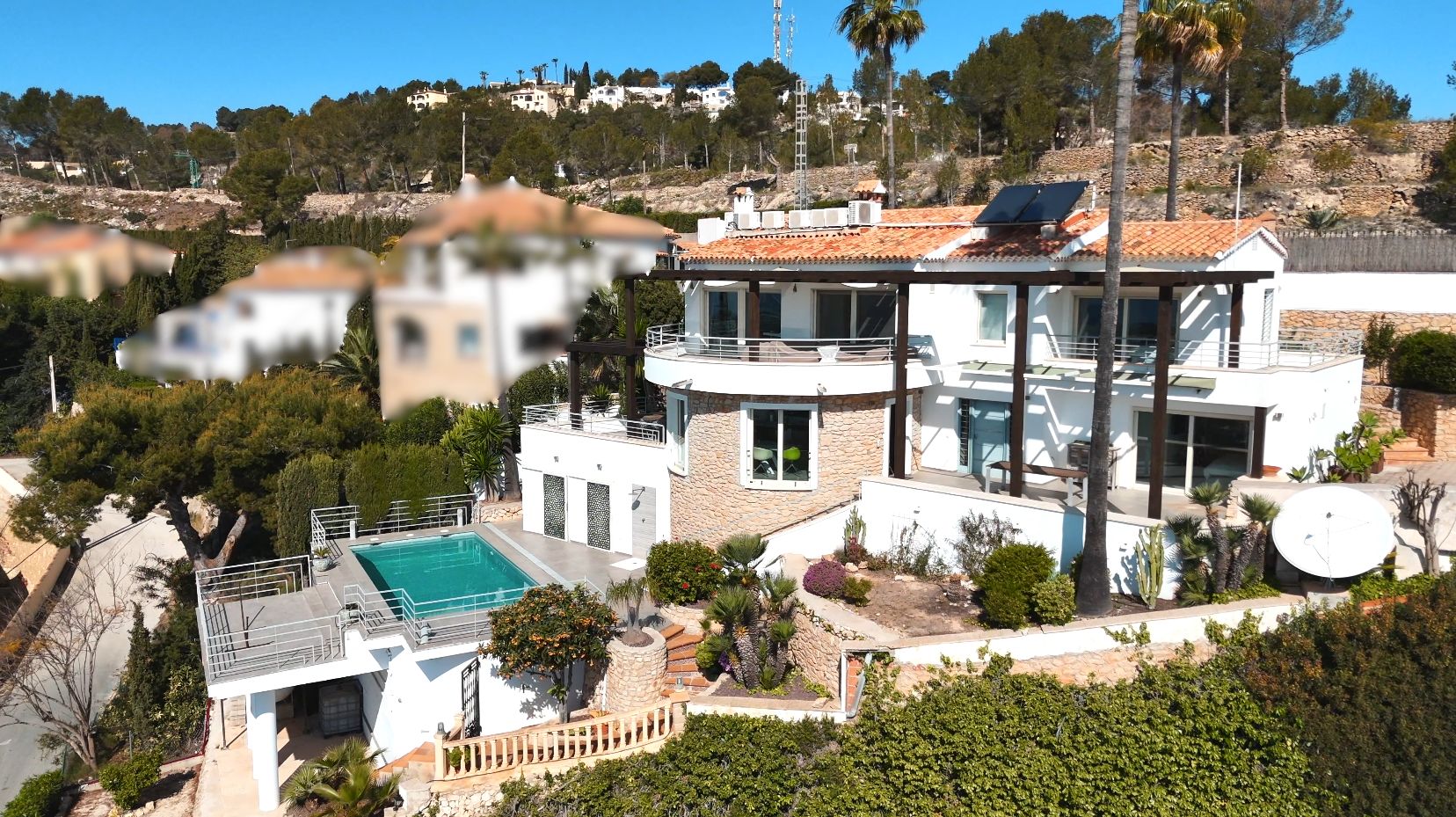 Mehrstöckige Villa mit weißer Fassade, Natursteinakzenten, Solaranlage und privatem Pool, eingebettet in eine mediterrane Gartenlandschaft mit Palmen.