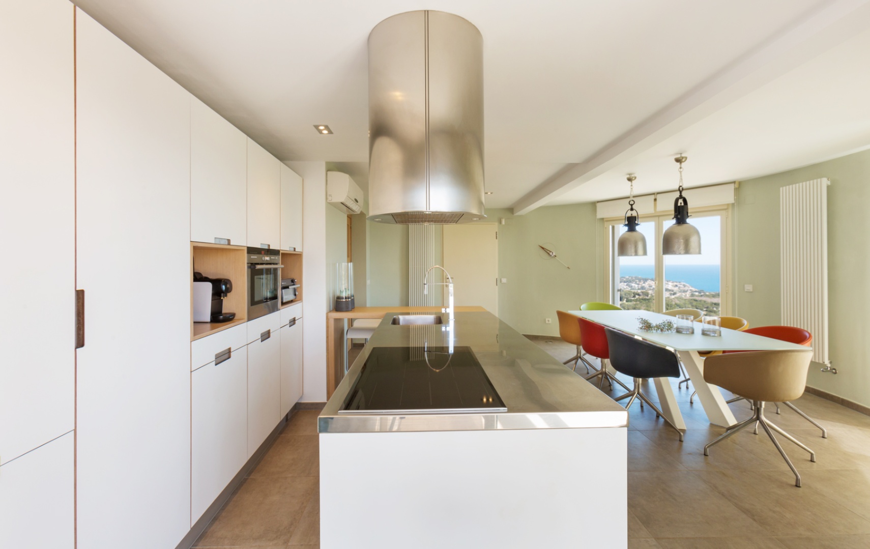 Moderne offene Küche mit weißer Kochinsel, Edelstahlarbeitsplatte, zylindrischer Dunstabzugshaube und Essbereich mit Meerblick durch große Glastüren.