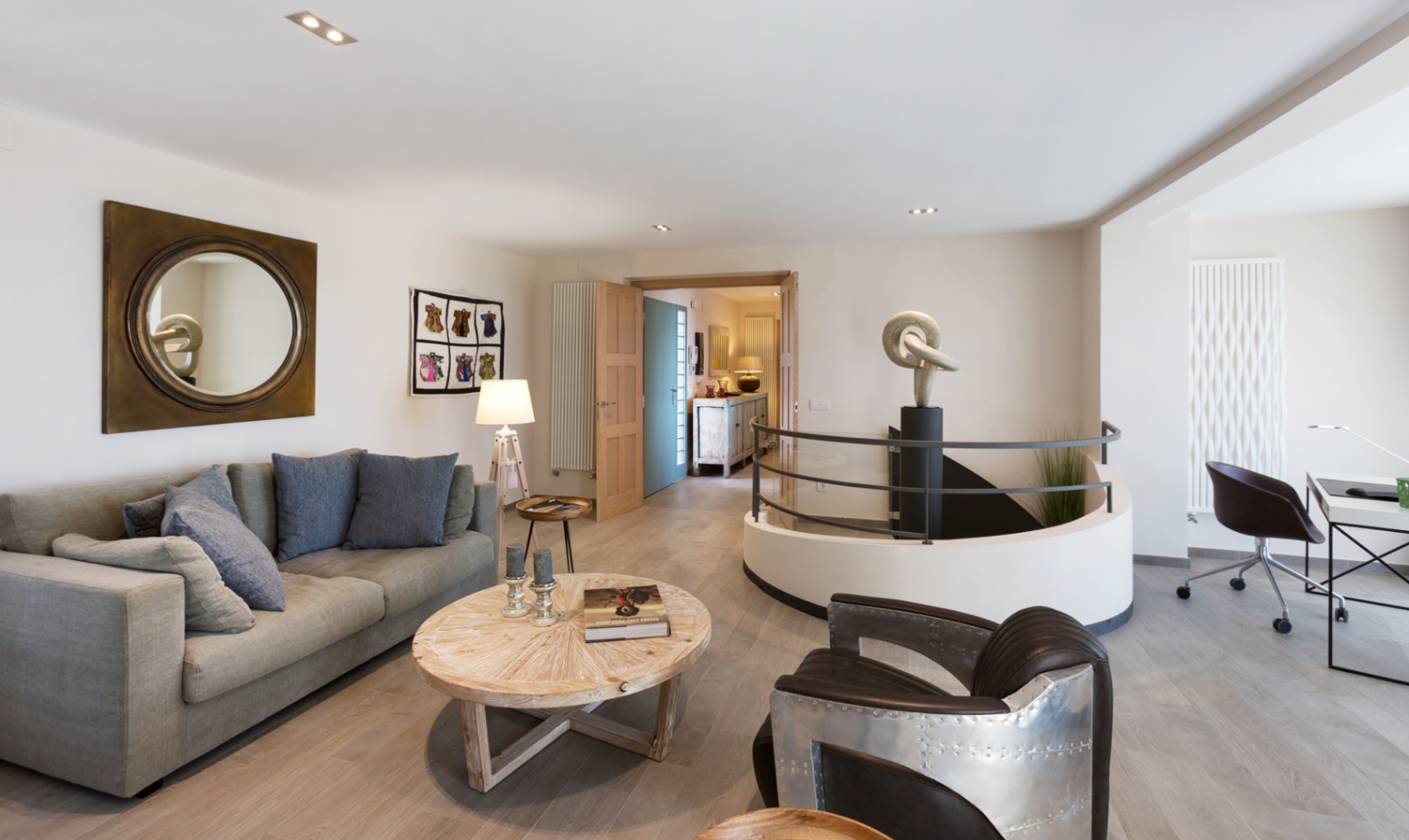 Moderner Wohnbereich mit grauem Sofa, Sessel im Aviator-Stil und runder Treppenöffnung mit Skulptur. Inklusive Arbeitsplatz und hellem Holzboden.