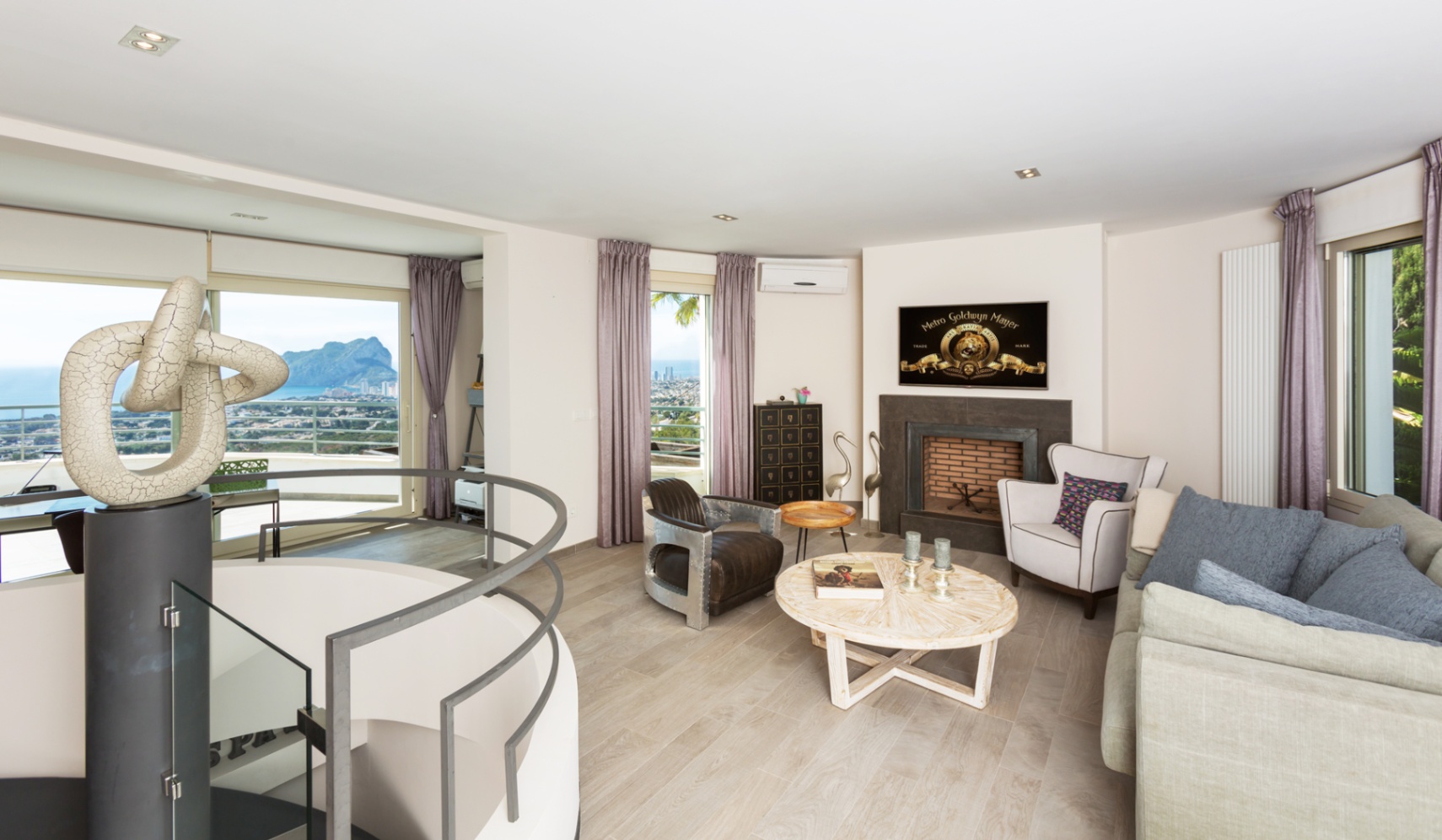 Moderner Wohnbereich mit Kamin, Wendeltreppe inklusive Skulptur und großen Fensterfronten mit Blick auf das Mittelmeer und den Peñón de Ifach.