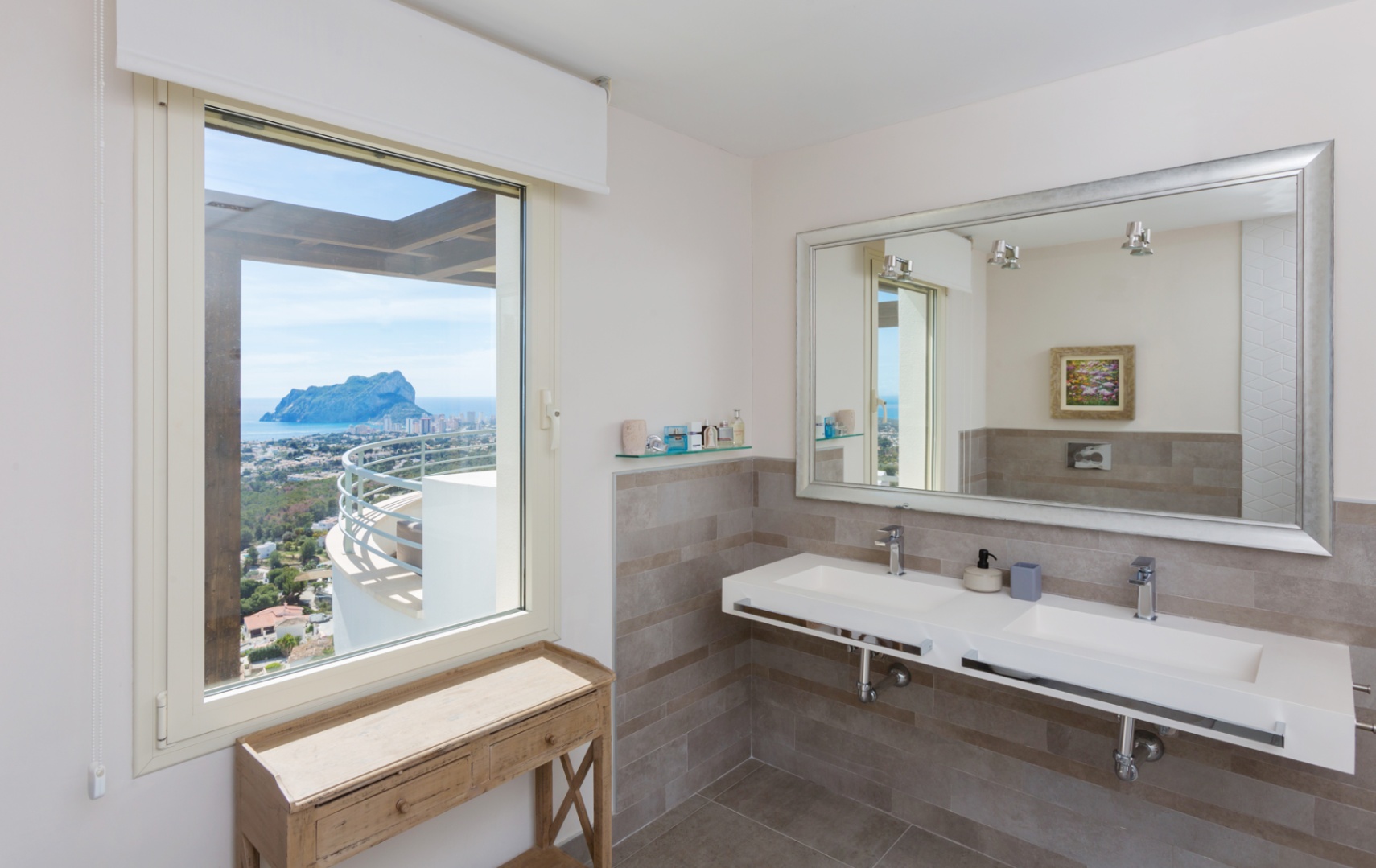 Modernes Badezimmer mit Doppel-Hängewaschbecken, großem Spiegel mit Silberrahmen und Fenster mit Panoramablick auf den Peñón de Ifach und das Mittelmeer.