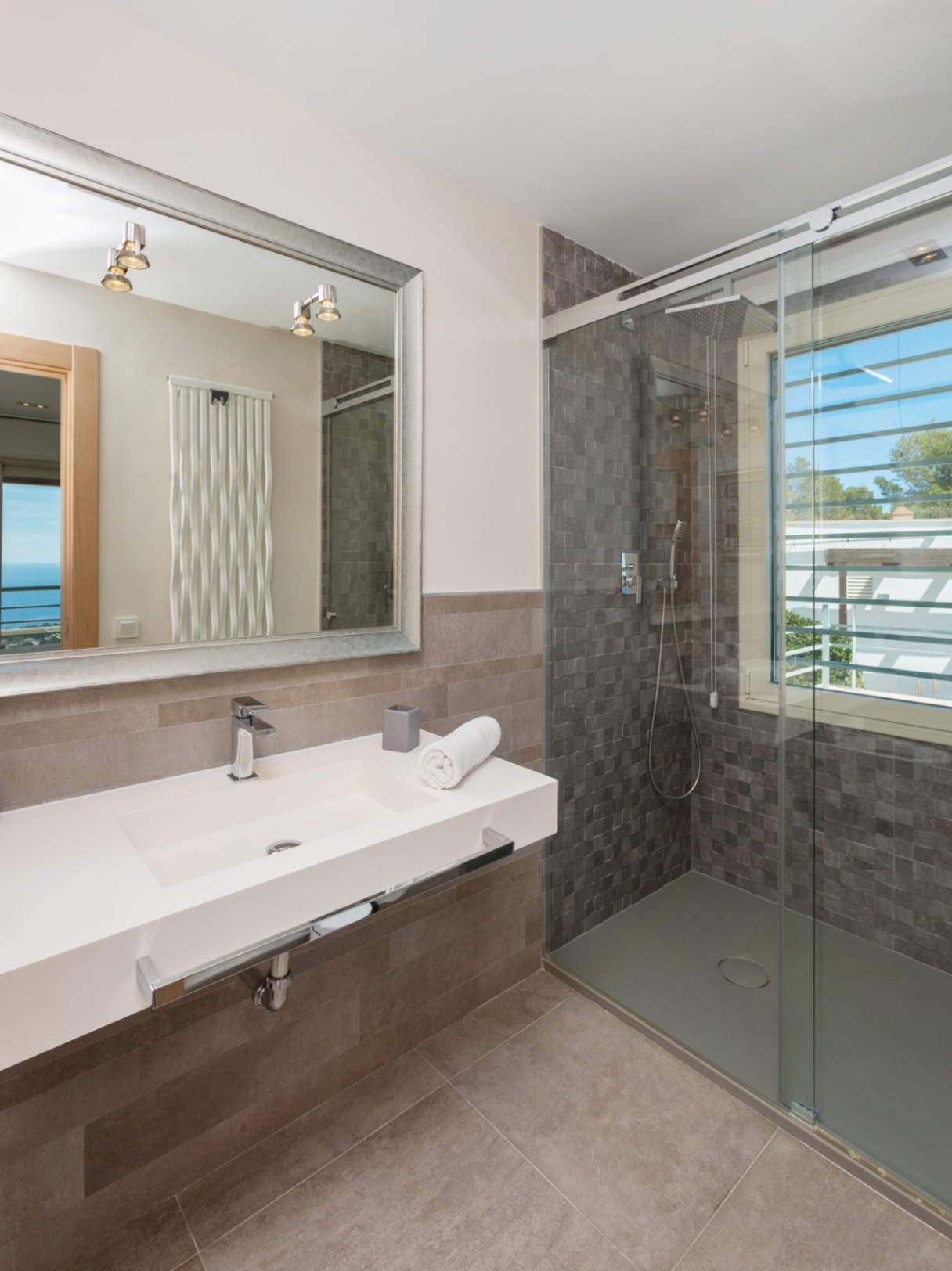 Modernes Badezimmer mit integriertem weißem Waschbecken, großem Spiegel mit Silberrahmen und begehbarer Dusche mit grauen Mosaikfliesen.