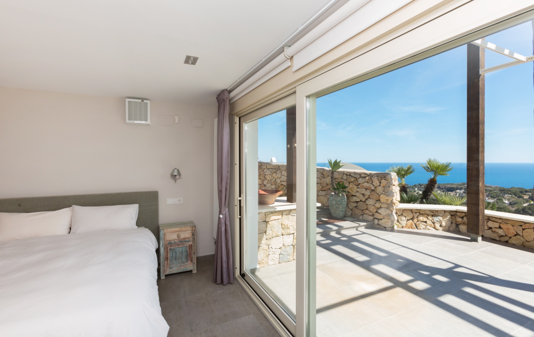 Schlafzimmer mit großen Glasschiebetüren, die auf eine Steinterrasse mit Panoramablick auf das Meer und rustikalem Nachttisch führen.