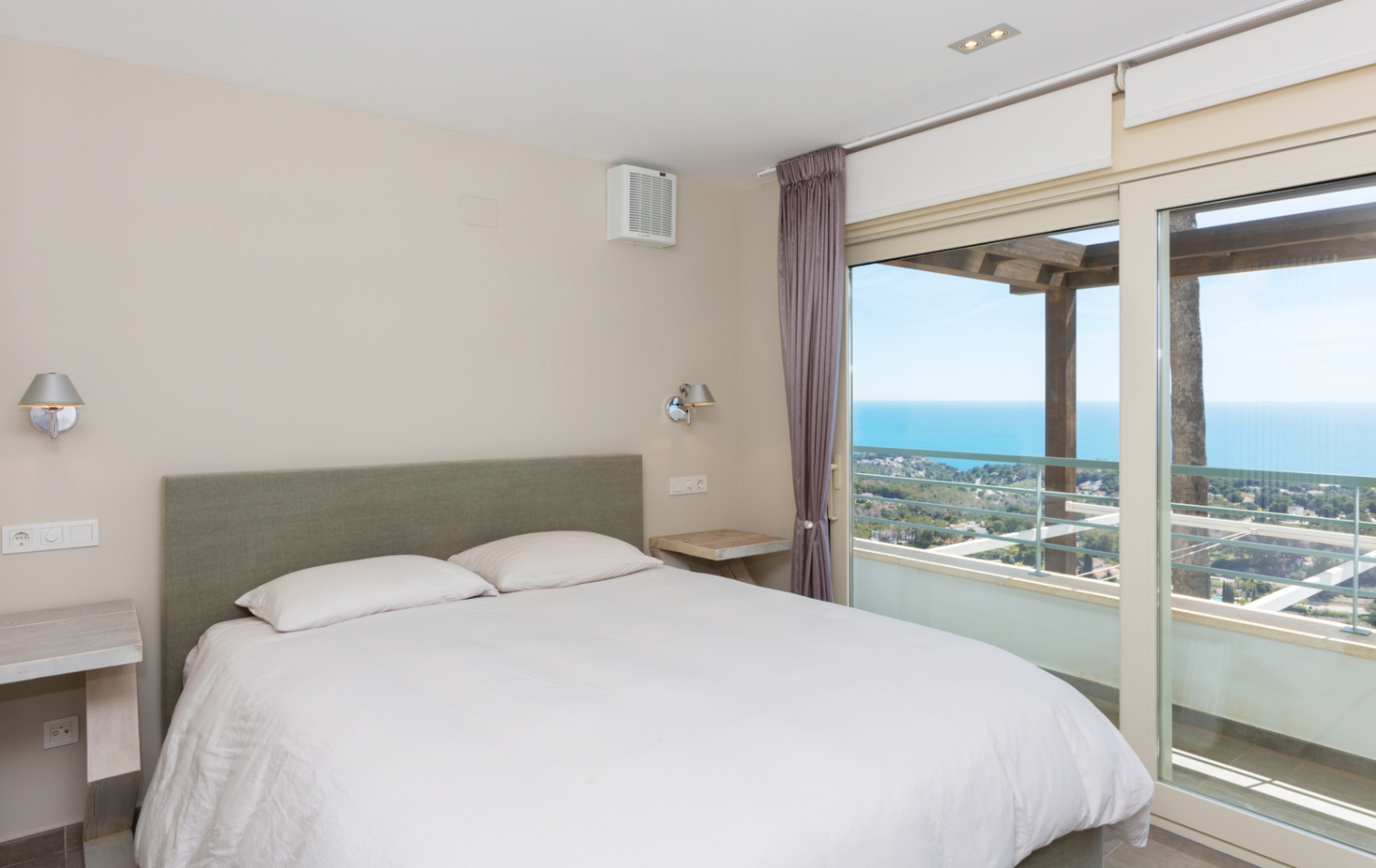 Schlafzimmer mit grün gepolstertem Kopfteil und großen Glasschiebetüren, die auf eine Terrasse mit Holzpergola und Panoramablick auf das Meer führen.
