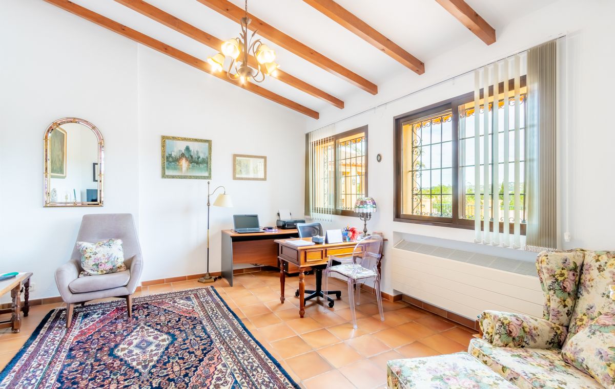 Arbeitszimmer mit sichtbaren Holzbalken, Terrakottaboden und zwei Arbeitsplätzen. Große Fenster mit Sicherheitsgittern sorgen für viel Tageslicht.