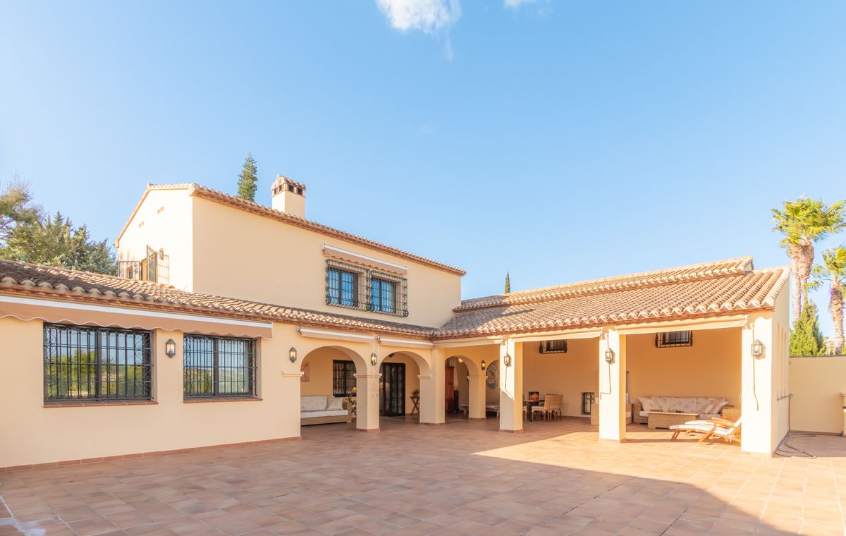 Außenansicht einer mediterranen Villa mit einer großen Terrasse aus Terrakottafliesen, Rundbogen-Veranden mit Sitzbereichen und cremefarbener Fassade.
