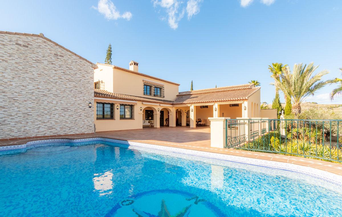 Außenansicht einer mediterranen Villa mit Mosaik-Pool, weitläufiger Terrakotta-Terrasse und überdachter Bogenveranda.