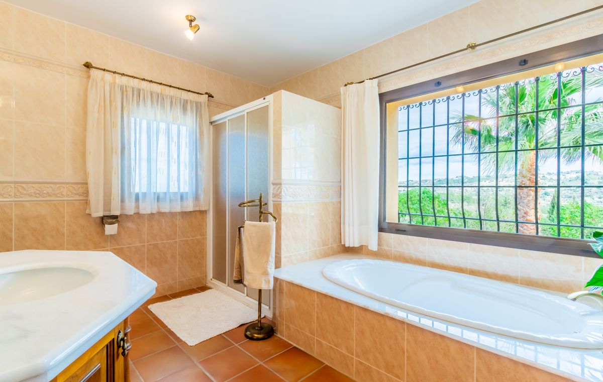 Badezimmer mit Einbauwanne unter dem Fenster, separater Duschkabine, weißem Marmorwaschtisch und rustikalen Terrakotta-Bodenfliesen.