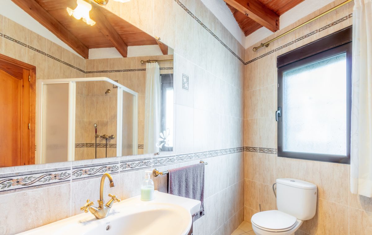 Badezimmer mit schräger Holzdecke, sichtbaren Balken, beigefarbenen Wandfliesen und klassischen Armaturen aus Messing.