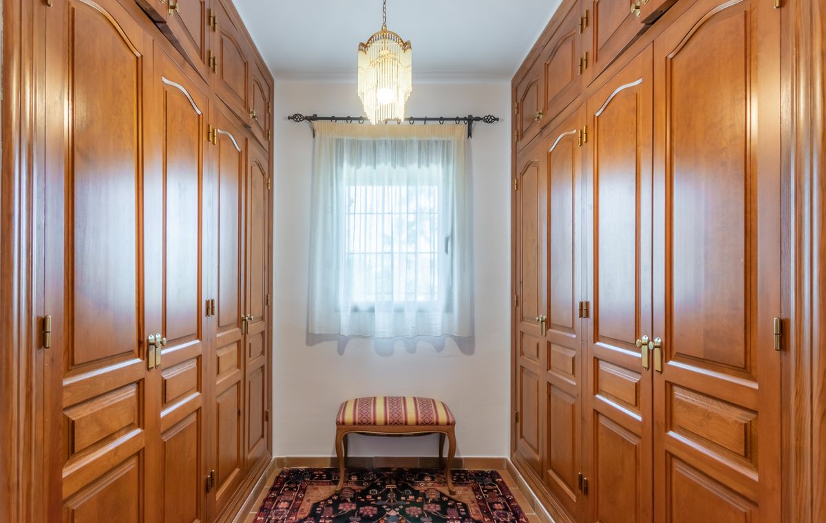 Begehbarer Kleiderschrank mit deckenhohen Holzschränken, klassischem Kronleuchter und zentralem Fenster für Tageslicht.