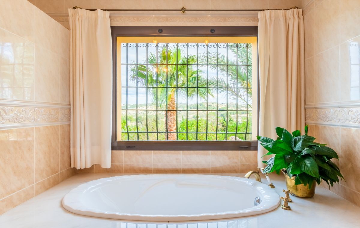 Eingelassene ovale Badewanne mit Messingarmaturen unter einem Fenster mit Schmiedeeisen und Blick auf Palmen und die grüne Landschaft.