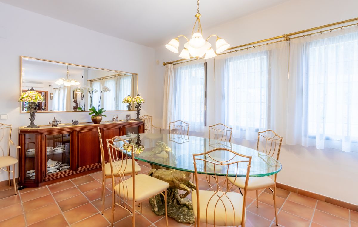 Formales Esszimmer mit ovalem Glastisch auf skulpturalem Messingfuß, goldfarbenen Stühlen, Terrakottaboden und einem Sideboard aus Holz mit Spiegel.