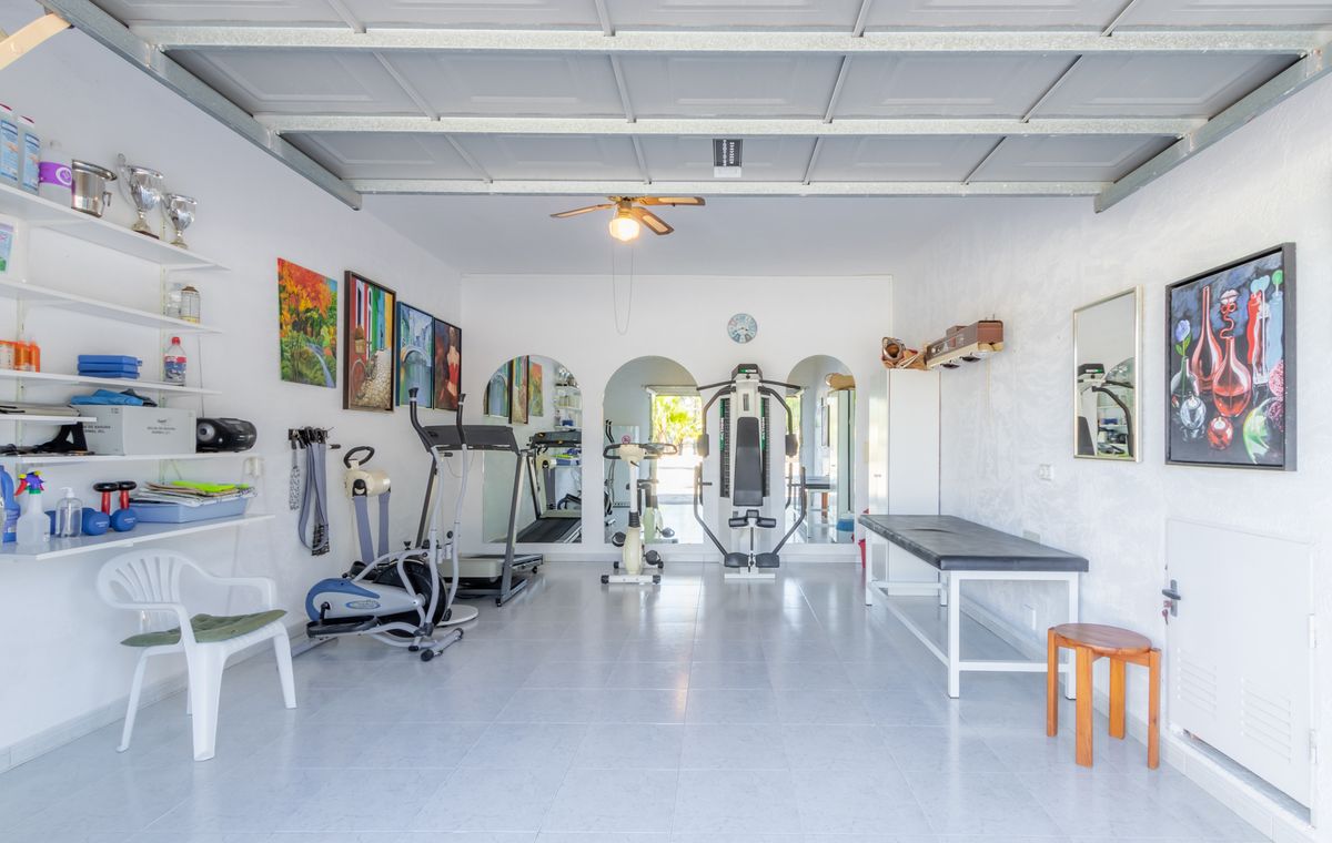 Privates Fitnessstudio in einer umgebauten Garage mit Fliesenboden, Kardiogeräten, einer Multistation und einer Massageliege unter einem Deckenventilator.