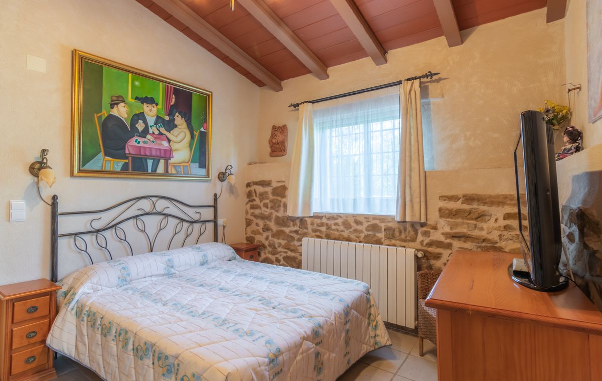 Rustikales Schlafzimmer mit Schmiedeeisenbett, Natursteinwänden und Holzbalkendecke. Ein großes figürliches Gemälde und eine Holzkommode mit Fernseher prägen den Raum.