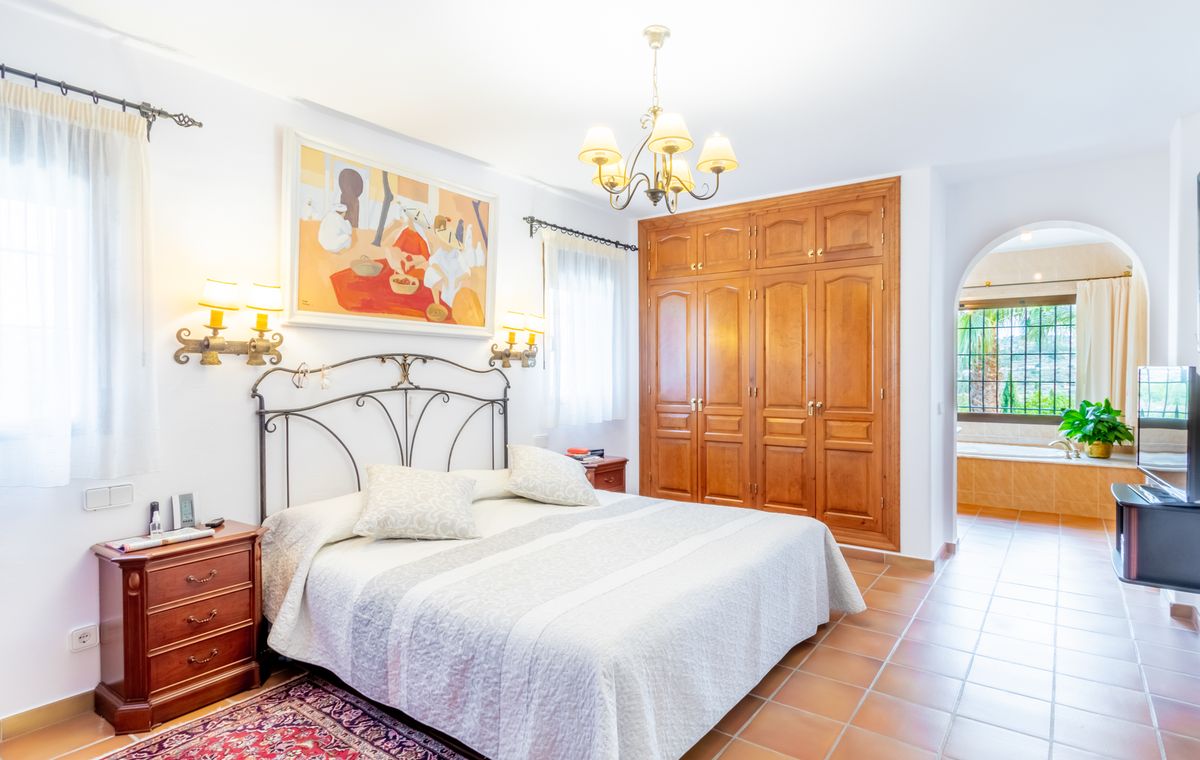 Schlafzimmer mit schmiedeeisernem Kopfteil, Einbauschränken aus Holz und Terrakottaboden. Ein Rundbogen führt zum En-Suite-Badezimmer mit Einbauwanne.
