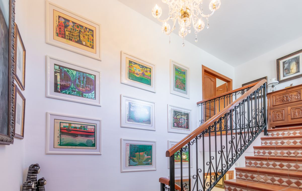 Treppenhaus mit Terrakotta-Stufen, schmiedeeisernem Geländer und einer Bildergalerie an der weißen Wand, ergänzt durch einen klassischen Kristallleuchter.