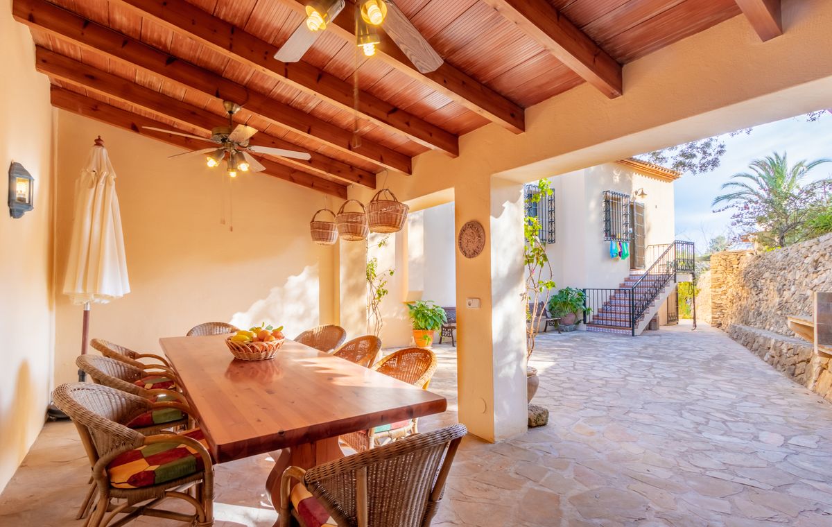 Überdachte Terrasse mit Holzbalkendecke, Deckenventilatoren und Essbereich aus Korbgeflecht, mit Blick auf einen gepflasterten Innenhof.