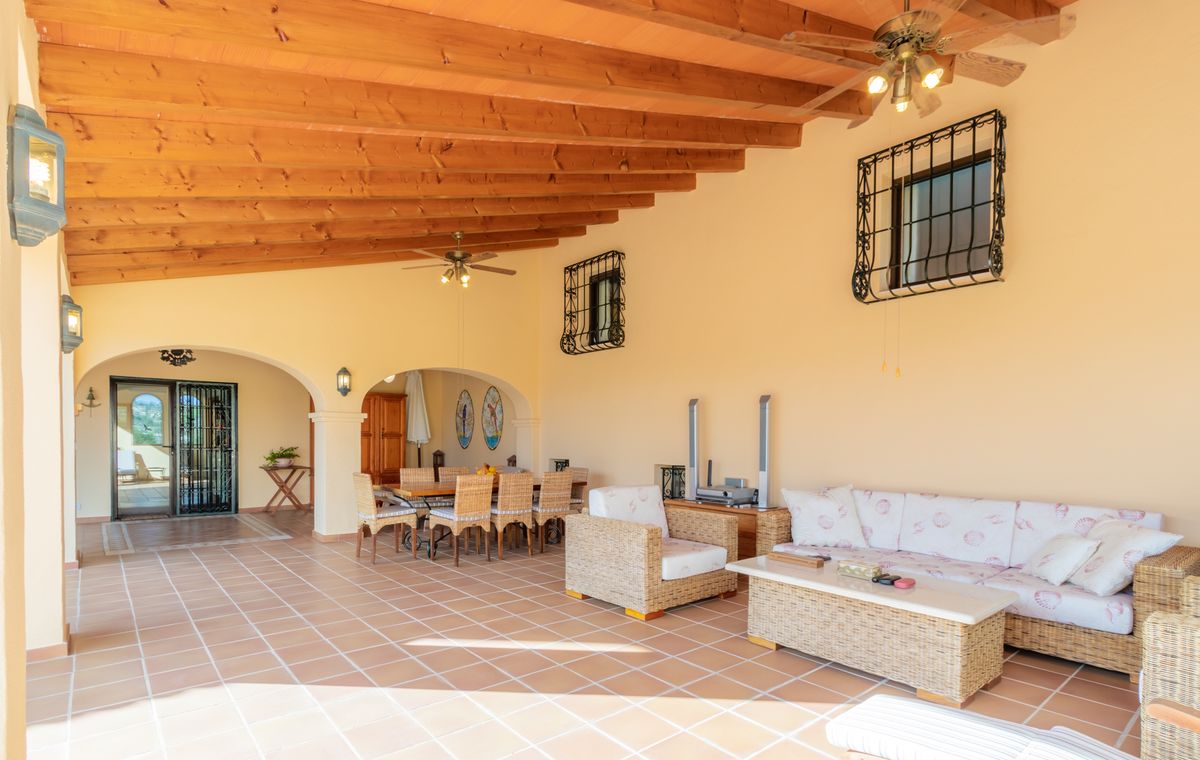 Überdachte Terrasse mit sichtbaren Holzbalken, Terrakottaboden und einem Loungebereich mit Korbmöbeln sowie einem Essplatz im Freien.