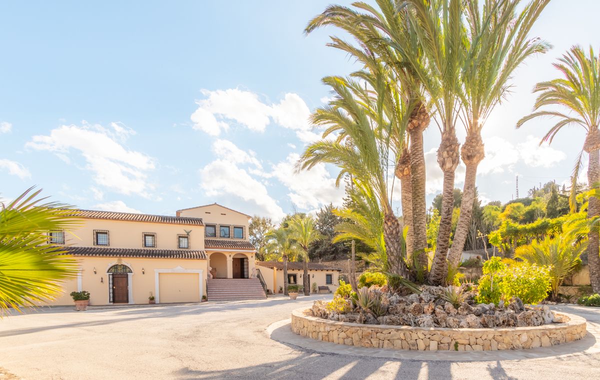 Weitläufige gepflasterte Einfahrt mit einem kreisförmigen Steinbeet und Palmen. Die Villa zeigt cremefarbene Fassaden und klassische mediterrane Ziegeldächer.