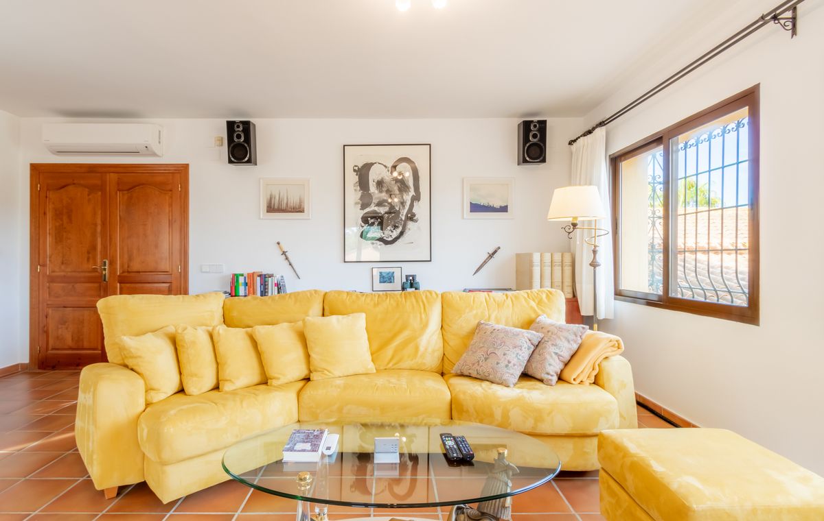 Wohnzimmer mit einem großen gelben Ecksofa, Glastisch und Terrakotta-Fliesenboden. Die weißen Wände sind mit Kunstwerken und integrierten Wandlautsprechern versehen.