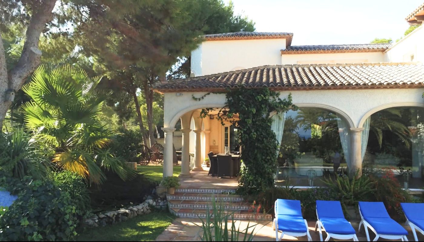Außenansicht einer mediterranen Villa mit bogenförmiger Veranda, Steinsäulen, Terrakottadach und einer Terrasse mit blauen Sonnenliegen.