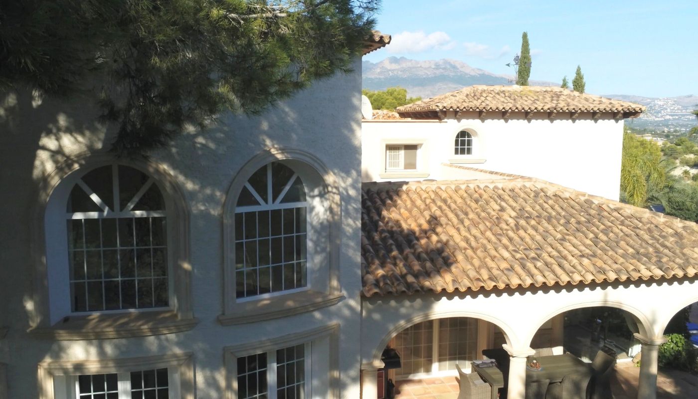 Außenansicht einer mediterranen Villa mit Terrakotta-Ziegeldächern, Rundbogenfenstern und überdachter Terrasse mit Blick auf die Berge.