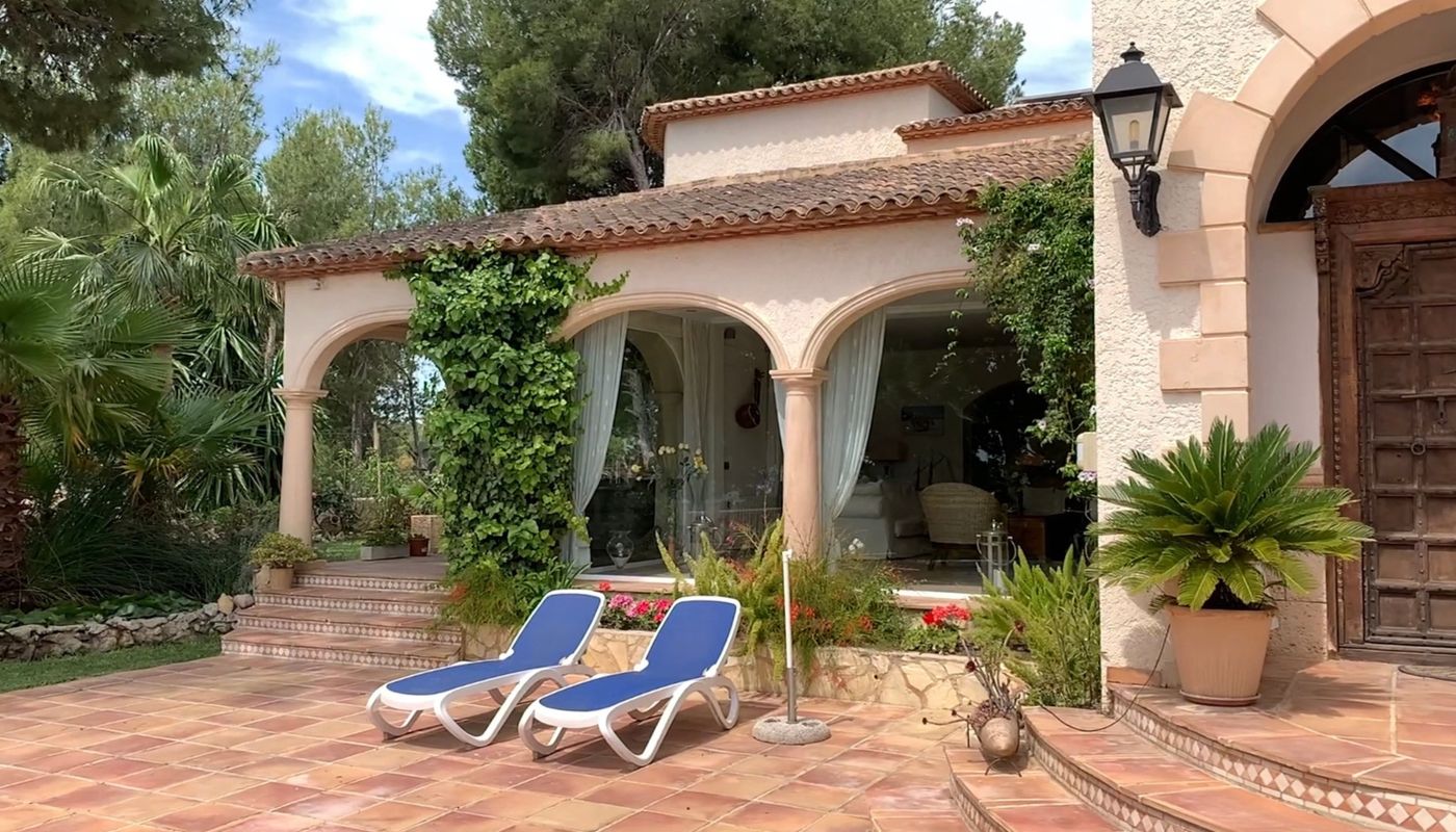 Außenliegende Terracotta-Terrasse mit blauen Sonnenliegen, einem bogenförmigen Säulengang mit Kletterpflanzen und einer massiven, geschnitzten Holztür.