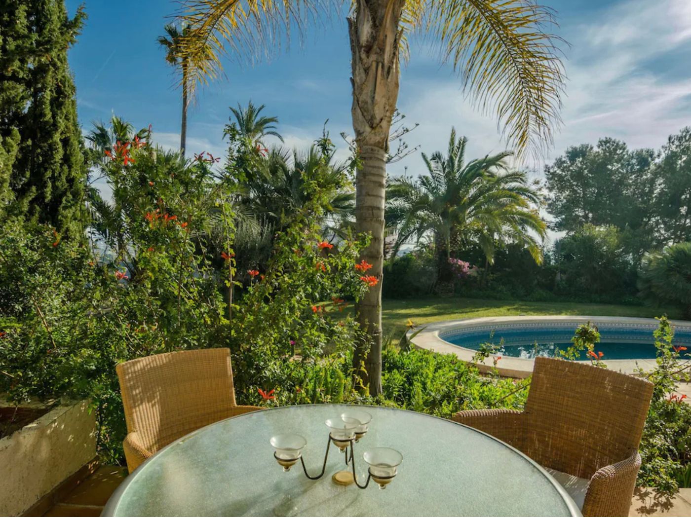 Außenterrasse mit Glastisch und Korbsesseln, mit Blick auf einen kreisförmigen Pool und üppige mediterrane Gärten mit ausgewachsenen Palmen.