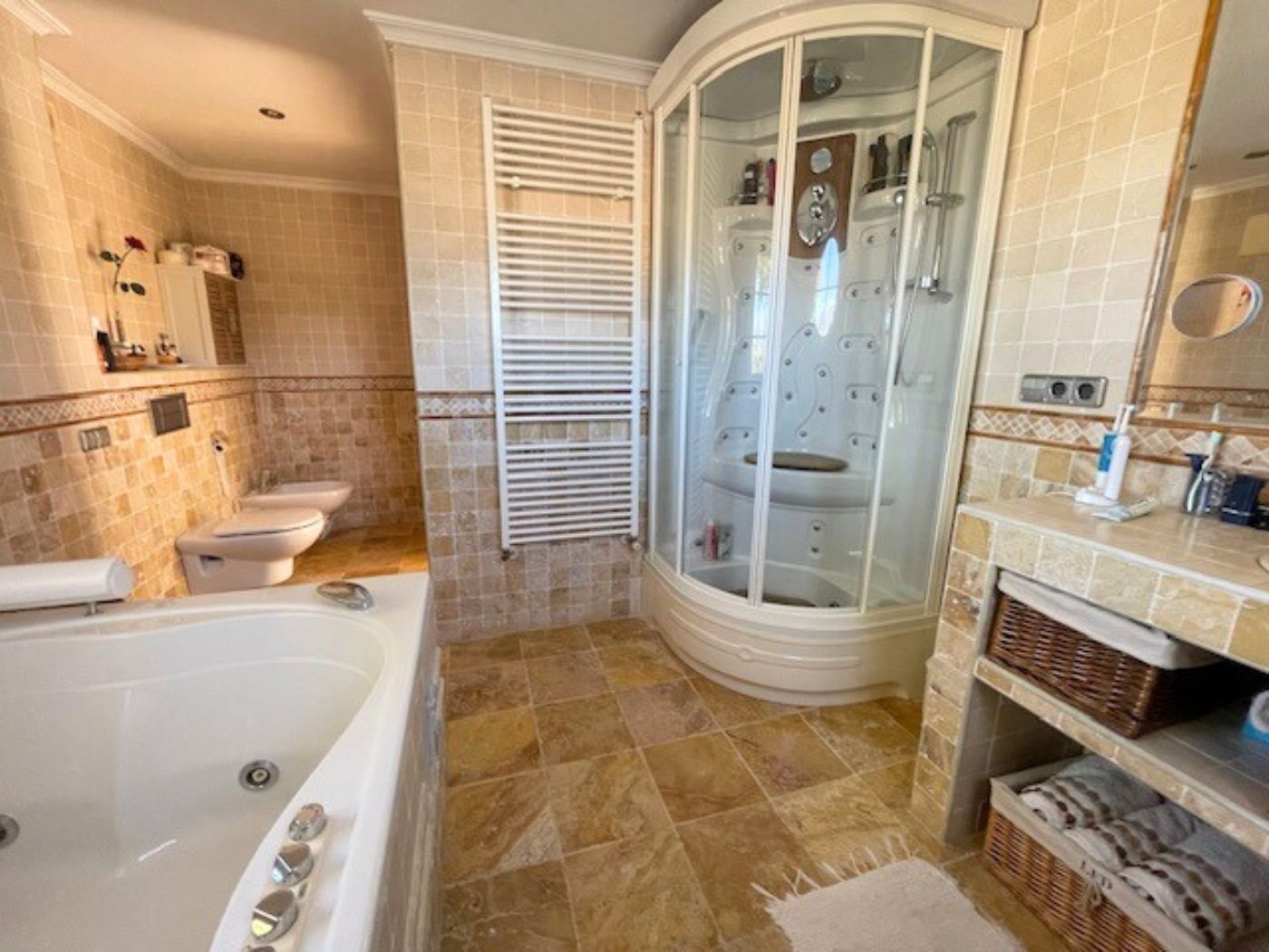 Badezimmer mit Eck-Hydromassage-Duschkabine, großer Badewanne und Natursteinfliesen. Ausgestattet mit einem Handtuchheizkörper und praktischen Flechtkörben.