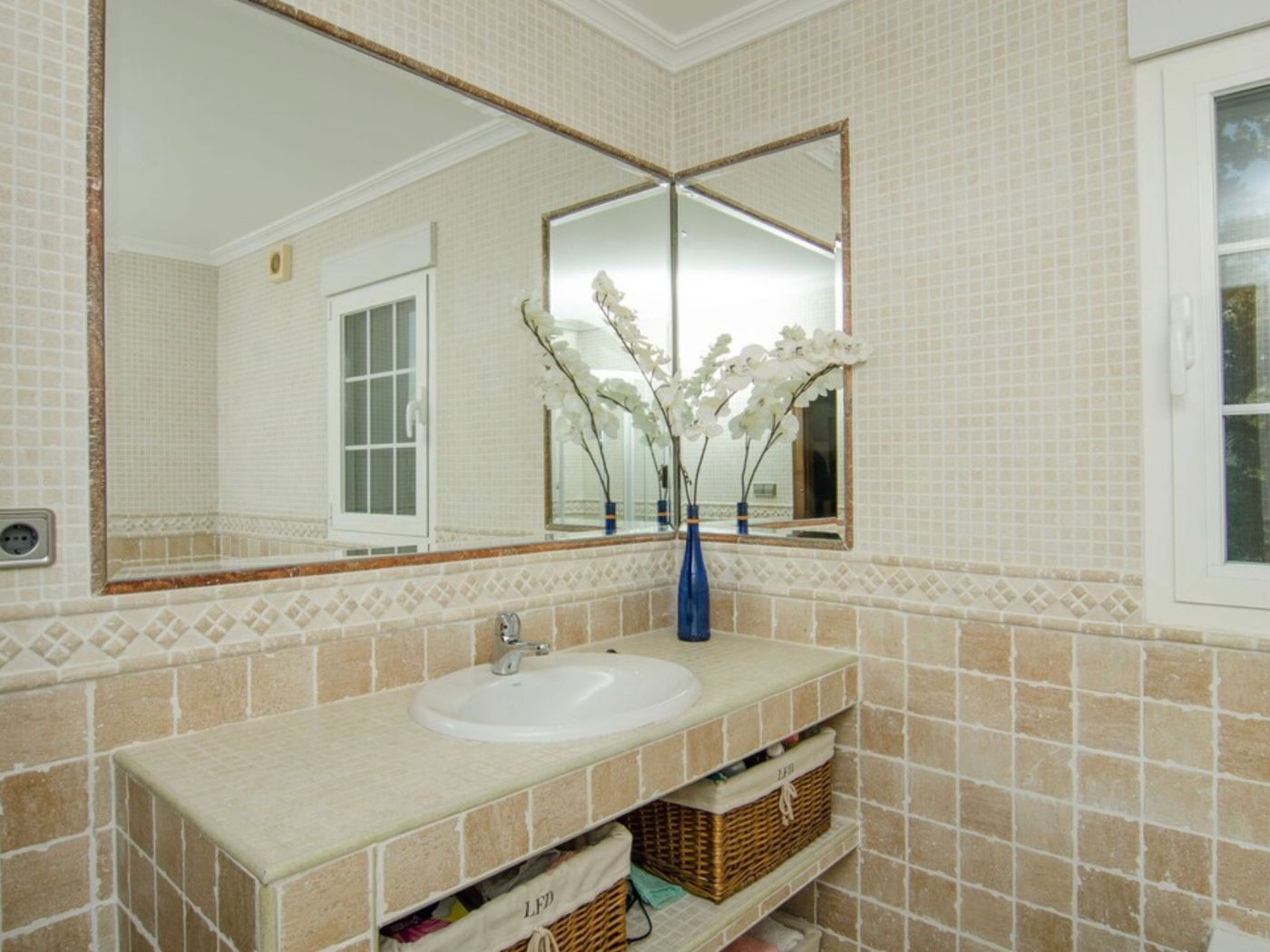 Badezimmer mit gefliestem Waschtisch, integriertem Waschbecken und Regalfächern mit Weidenkörben. Große Eckspiegel betonen die hellen Mosaikwände.