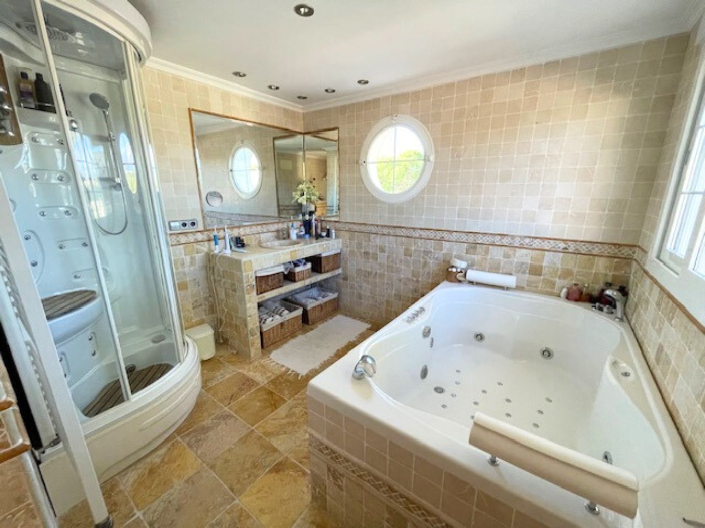 Badezimmer mit Hydromassage-Badewanne, Eckduschkabine und hellen Fliesenwänden, ergänzt durch ein charakteristisches Rundfenster.