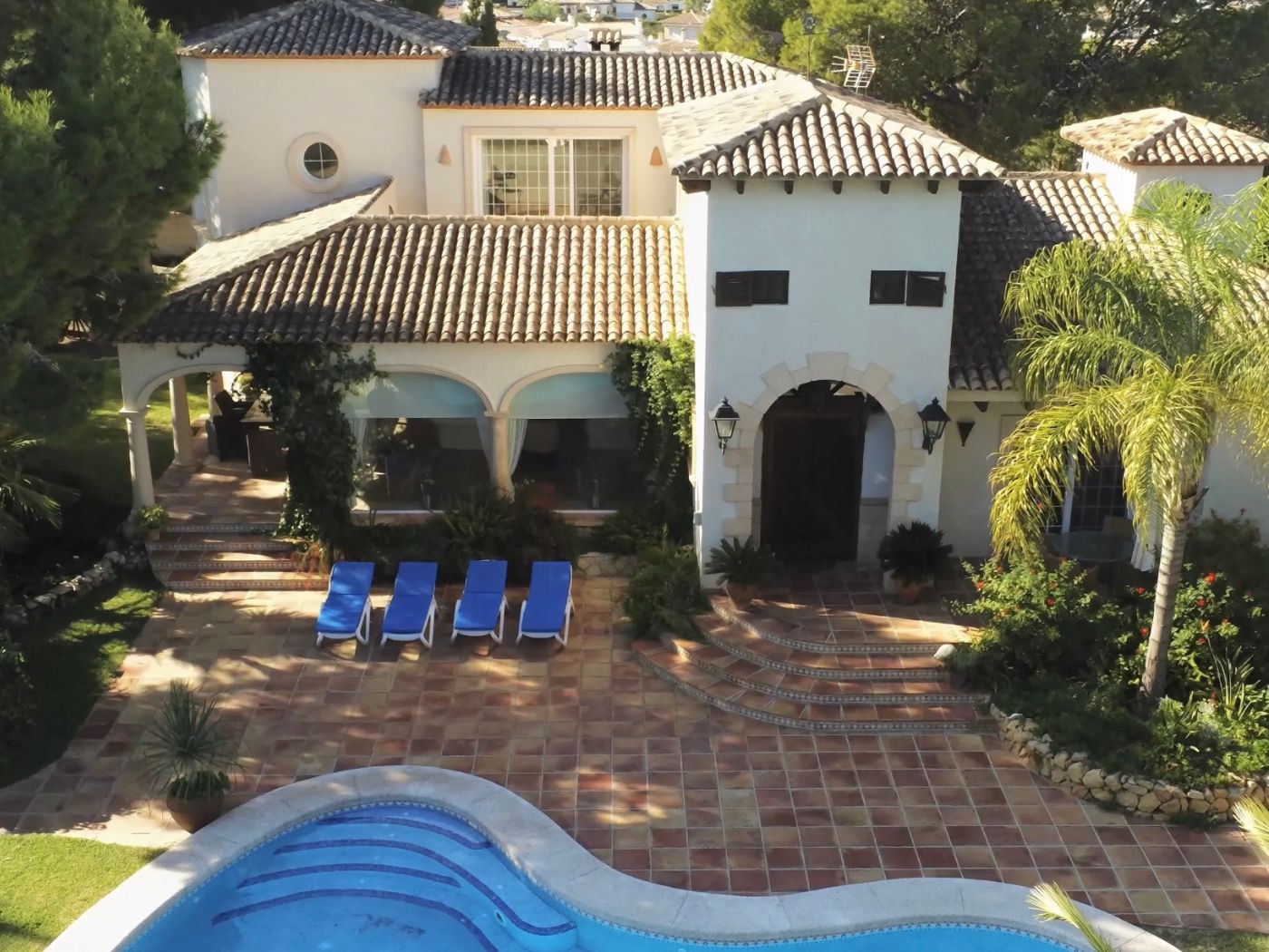 Erhöhte Ansicht einer mediterranen Villa mit geschwungenem Pool, Terrakotta-Terrasse mit vier Sonnenliegen und weißer Fassade mit steinernen Rundbogen-Eingängen.