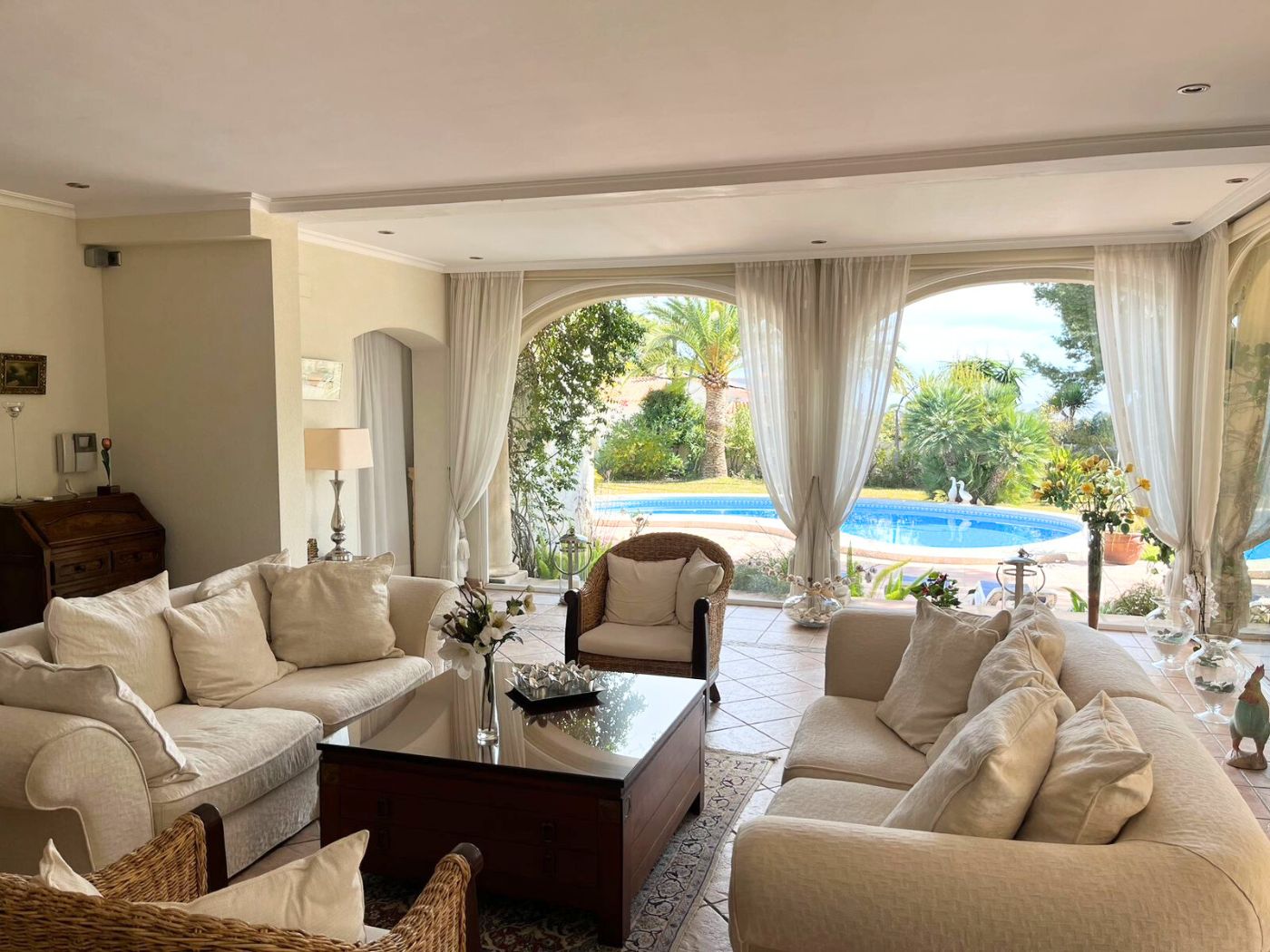 Großzügiges Wohnzimmer mit cremefarbenen Sofas und Korbsesseln, dessen große Rundbogenfenster einen direkten Blick auf den Pool und den Garten bieten.