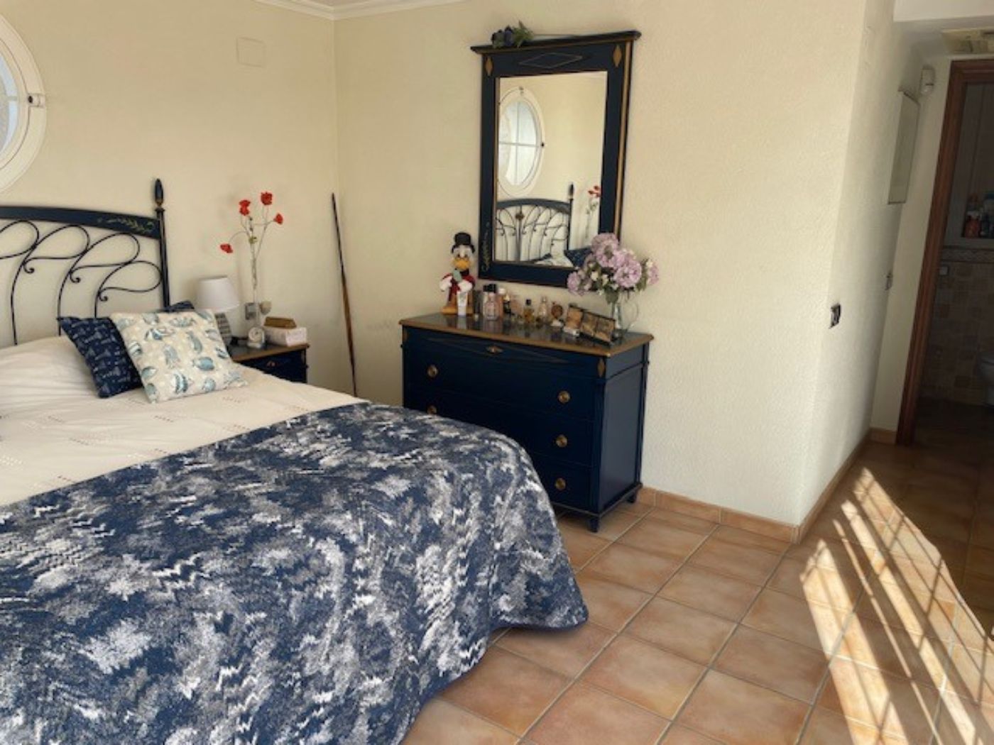 Schlafzimmer mit schmiedeeisernem Bettgestell, einer dunkelblauen Kommode mit passendem Spiegel und traditionellen Terrakotta-Fliesen sowie Rundfenster.