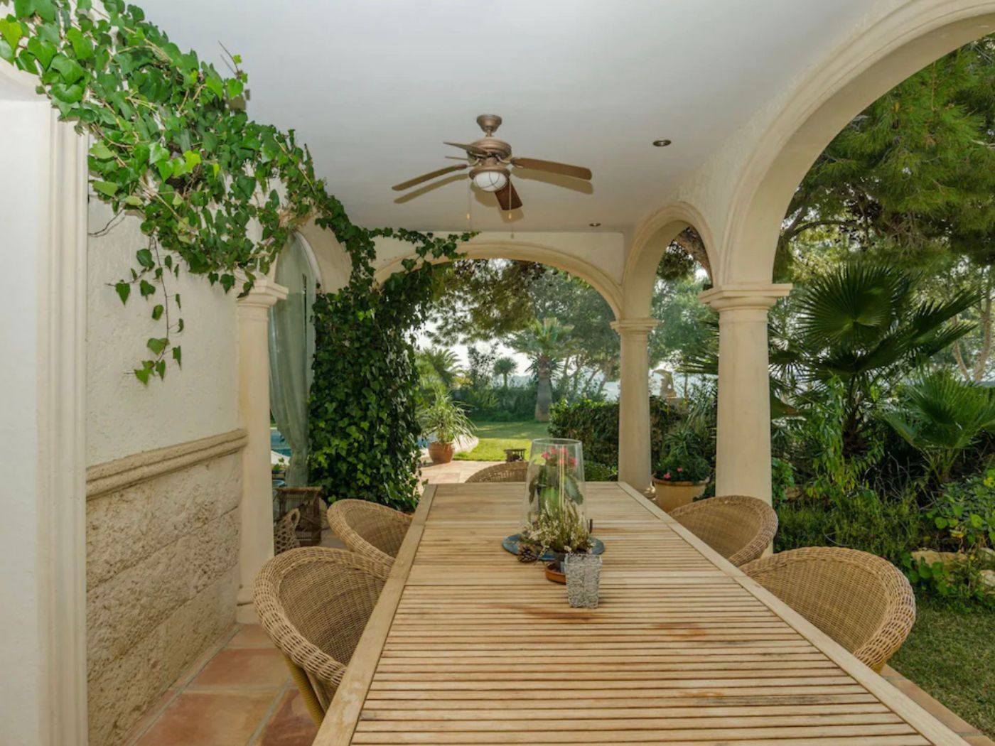Überdachte Terrasse mit klassischen Rundbögen, Holzesstisch und Korbstühlen, umgeben von Kletterpflanzen und Blick auf den mediterranen Garten.