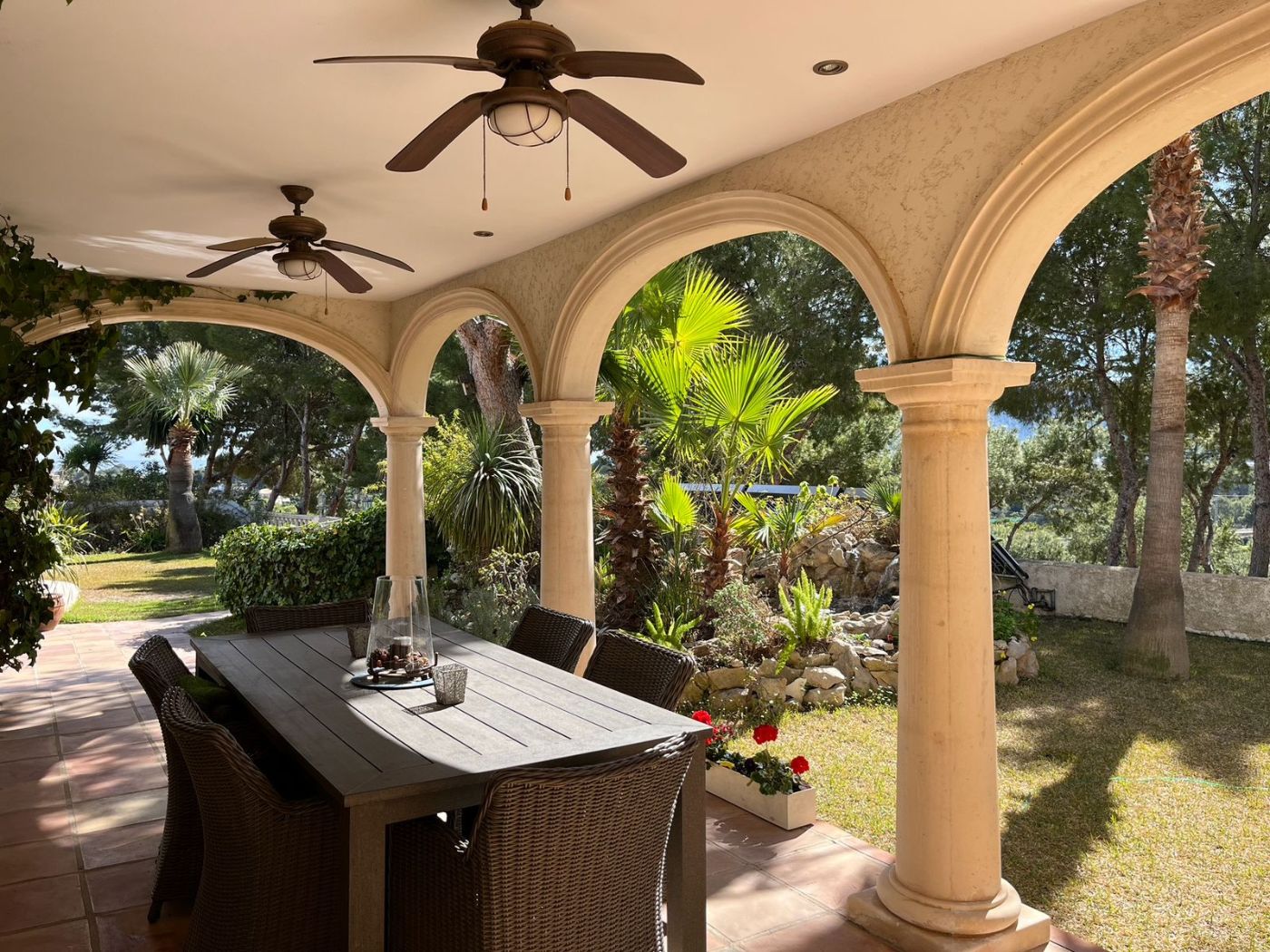 Überdachte Terrasse mit steinernen Rundbögen, Terrakottaboden und Essbereich im Freien mit Blick auf einen mediterranen Garten mit Palmen.