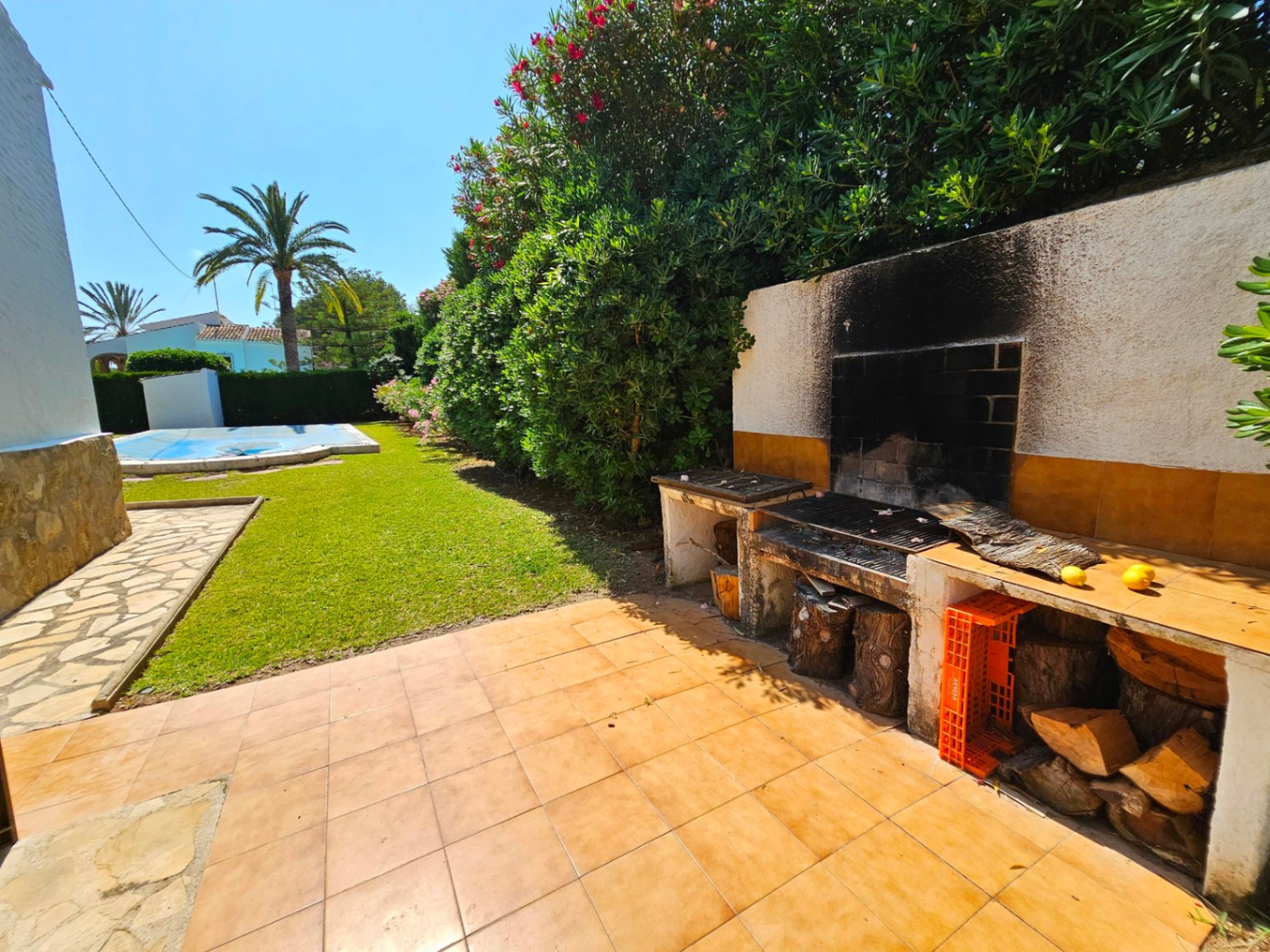 Außen-Grillstation mit Terrakotta-Fliesen und Holzlager, mit Blick auf einen gepflegten Rasen, den abgedeckten Pool und Palmen.