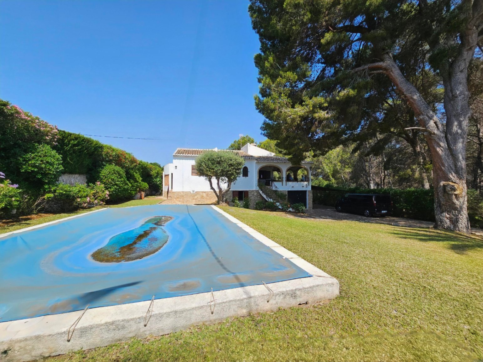 Außenansicht einer weißen Villa mit Bogenterrasse und großer Rasenfläche mit abgedecktem Pool, umgeben von alten Pinien und mediterraner Vegetation.