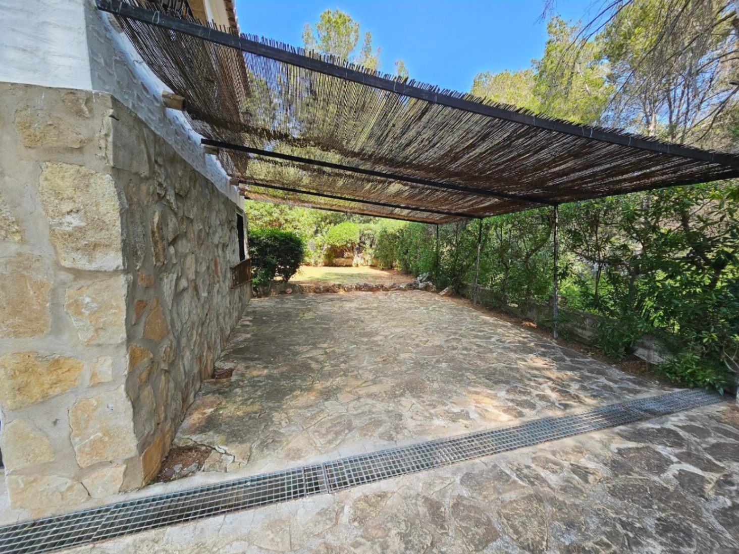 Außenbereich mit Natursteinpflaster und einer rustikalen Schilfrohr-Pergola, angrenzend an eine Steinmauer und umgeben von mediterraner Bepflanzung.