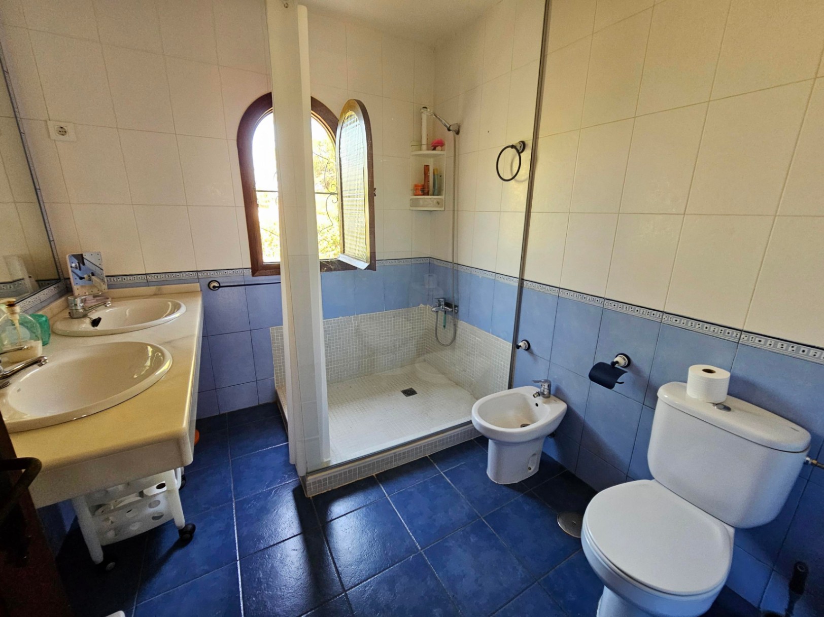 Badezimmer mit Doppelwaschtisch, Mosaikdusche und Rundbogenfenster. Blaue und weiße Fliesen an Wänden und Boden bieten natürliches Licht.