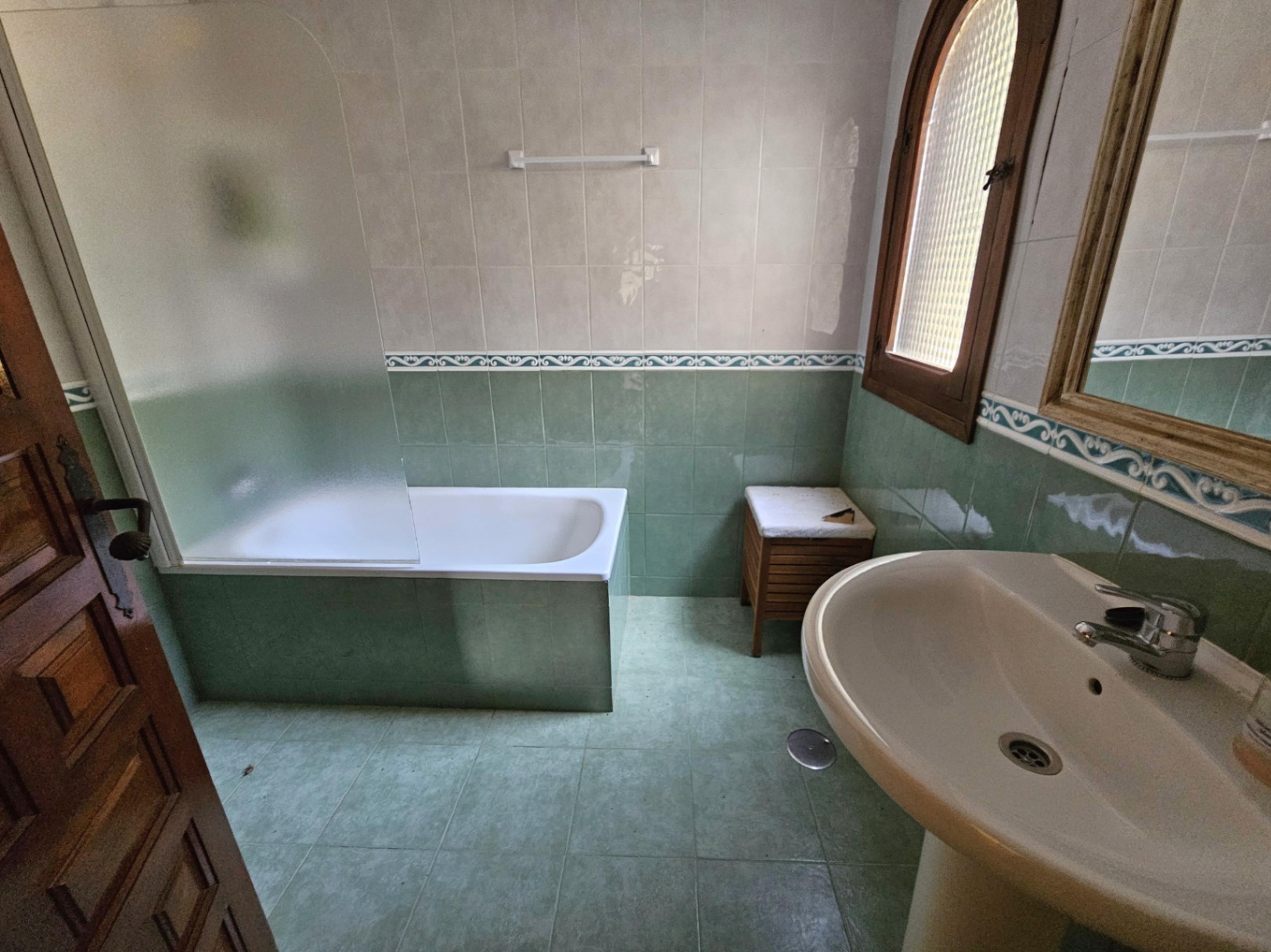 Badezimmer mit Einbaubadewanne, satinierter Glastrennwand, grünen Fliesen mit Zierbordüre und einem Rundbogenfenster aus Holz.