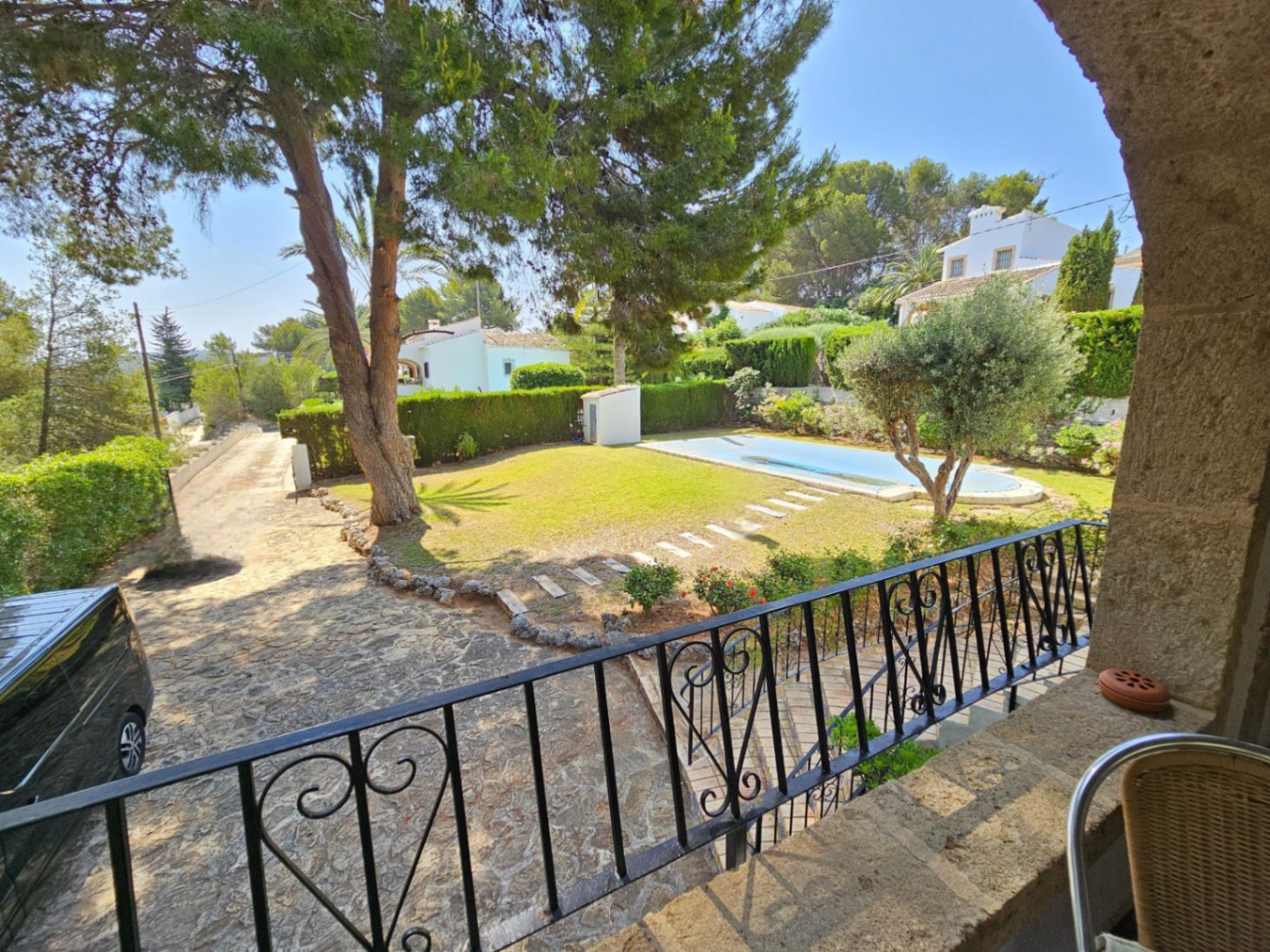 Blick von einem Steinbalkon mit Schmiedeeisengeländer auf einen Garten mit Pool, alten Pinien und einer gepflasterten Einfahrt.