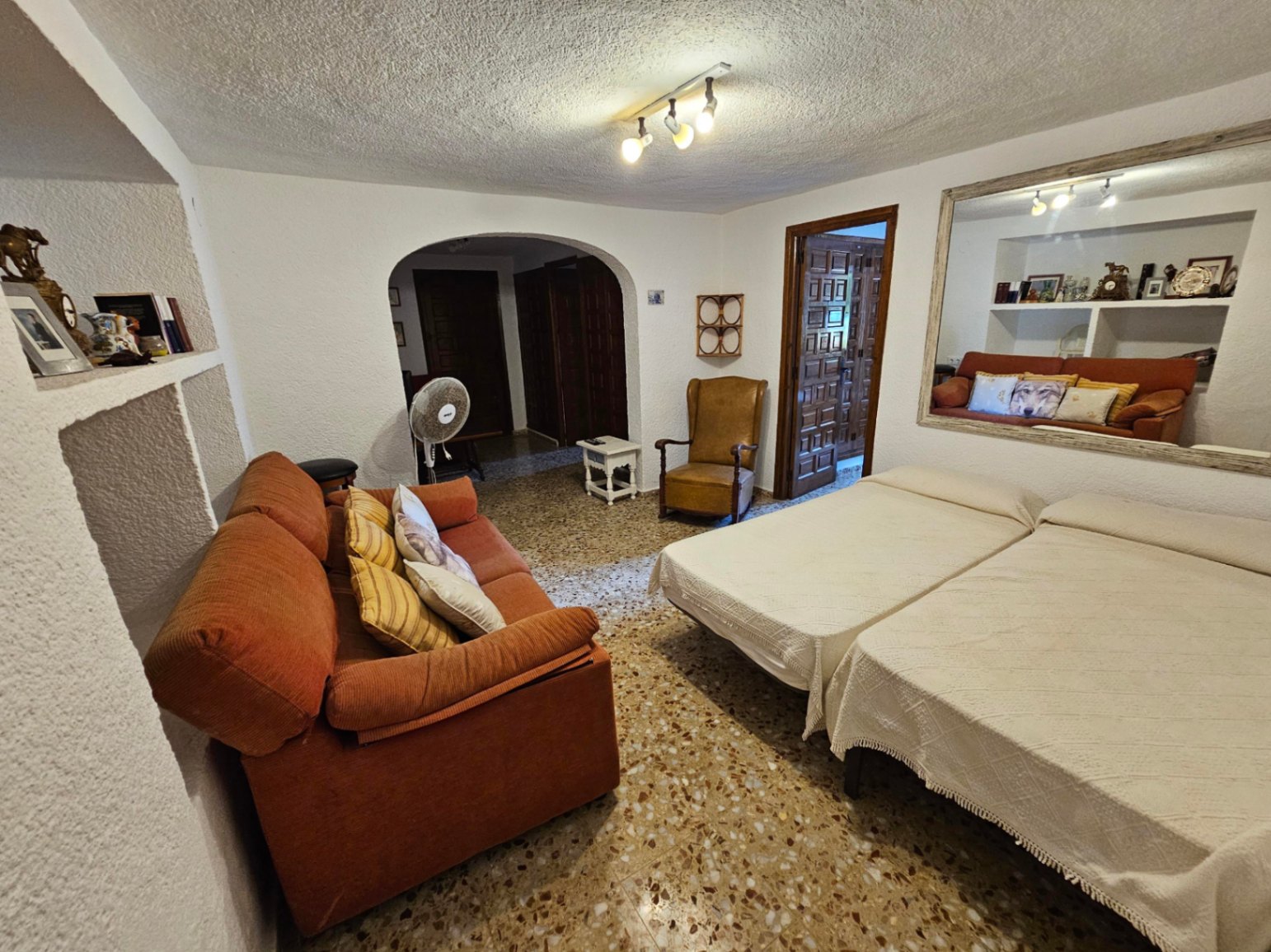 Mehrzweckraum mit zwei Einzelbetten, orangefarbenem Sofa und großem Spiegel. Mit Terrazzoboden, strukturierten Wänden und einem Rundbogen zum Flur.