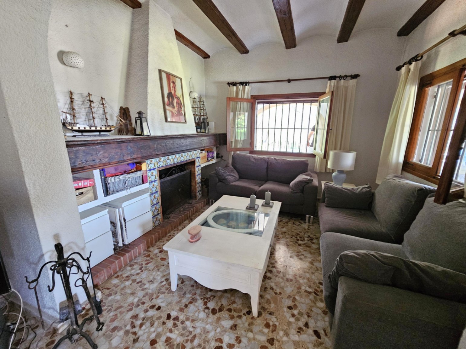 Rustikales Wohnzimmer mit Kamin, verzierten Fliesen und massiven Deckenbalken. Der Raum besticht durch traditionellen Terrazzoboden und integrierte Regalelemente.