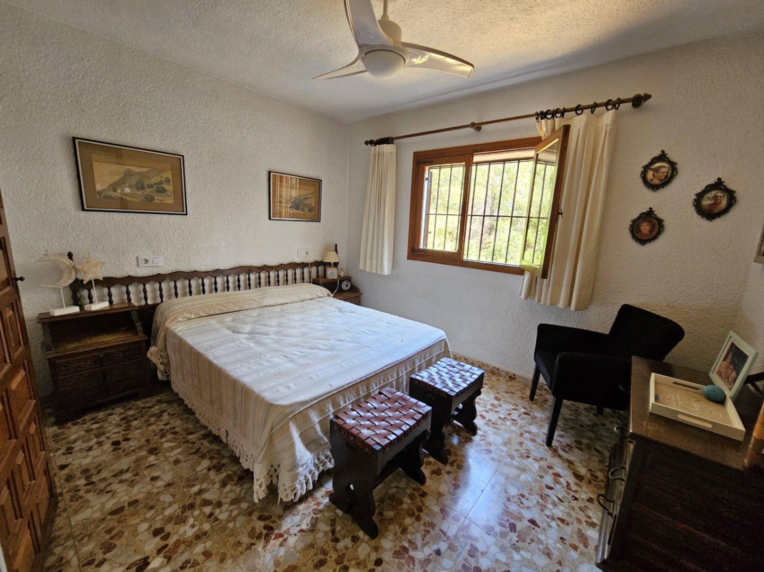 Schlafzimmer mit traditionellem Terrazzoboden, gedrechseltem Holzkopfteil und Lederhockern. Ein weißer Deckenventilator und ein Holzfenster sorgen für Helligkeit.