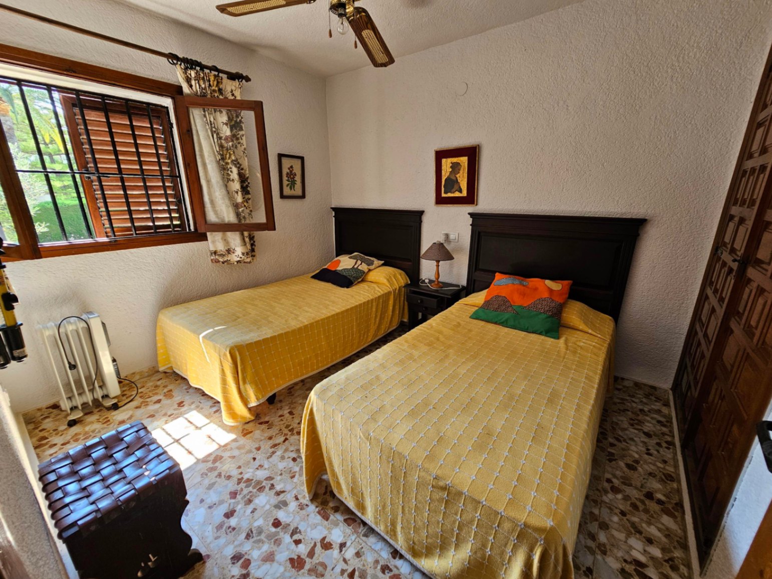 Schlafzimmer mit zwei Einzelbetten, dunklen Holzkopfteilen und klassischem Terrazzoboden. Mit Deckenventilator und Fenster mit Sicherheitsgittern.
