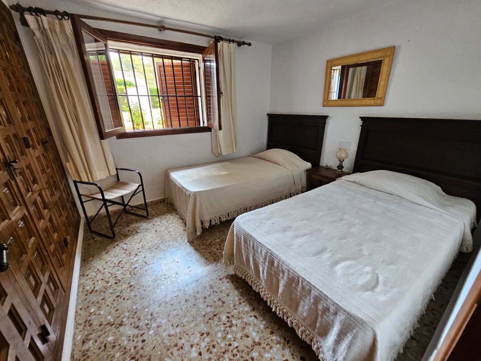 Schlafzimmer mit zwei Einzelbetten, dunklen Holzkopfteilen und Terrazzoboden. Mit eingebautem Holzschrank und Fenster mit Sicherheitsgittern und Schlagläden.