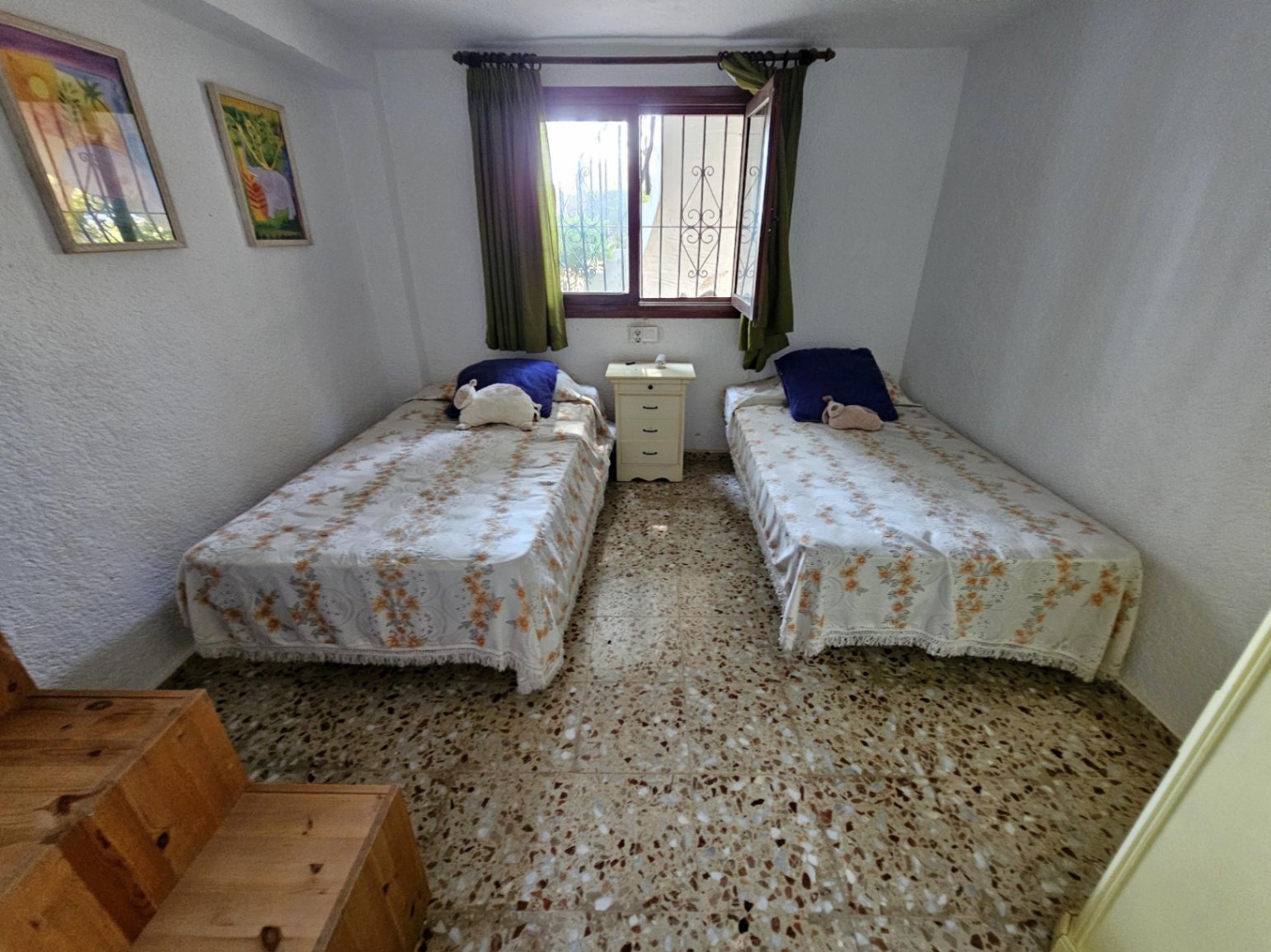 Schlafzimmer mit zwei Einzelbetten, Terrazzoboden und einem Fenster mit Sicherheitsgittern und grünen Vorhängen. Ein weißer Nachttisch steht zwischen den Betten.
